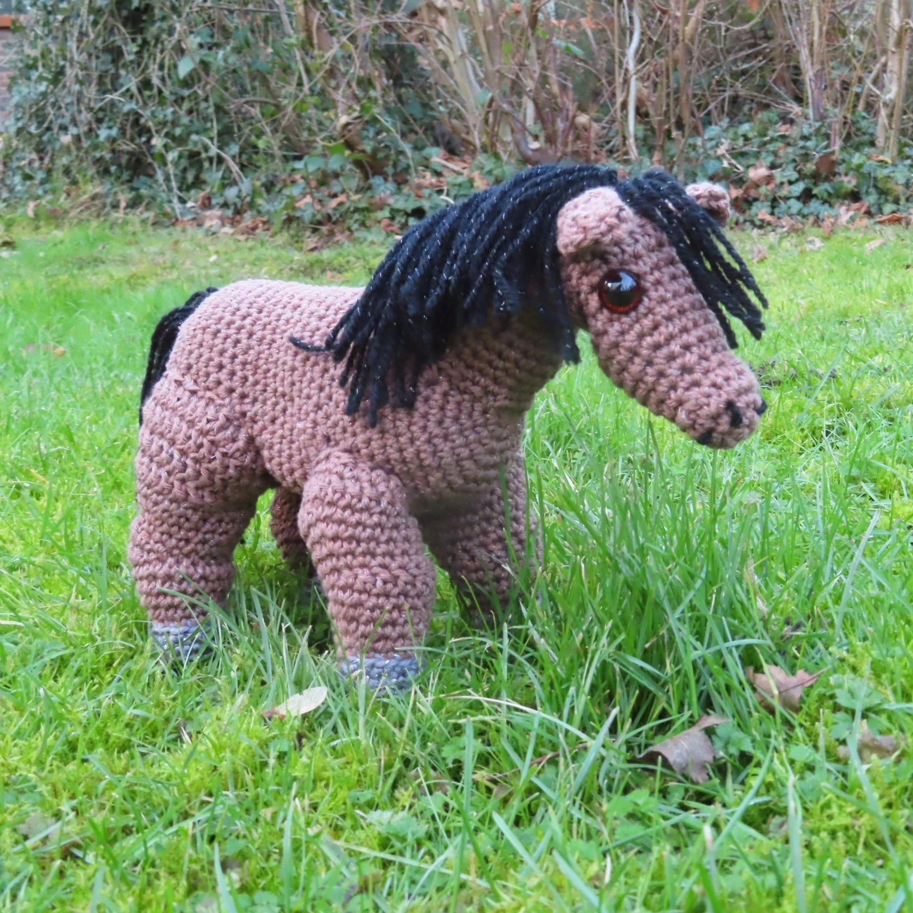 A brown crocheted horse with a black mane on a grassy lawn