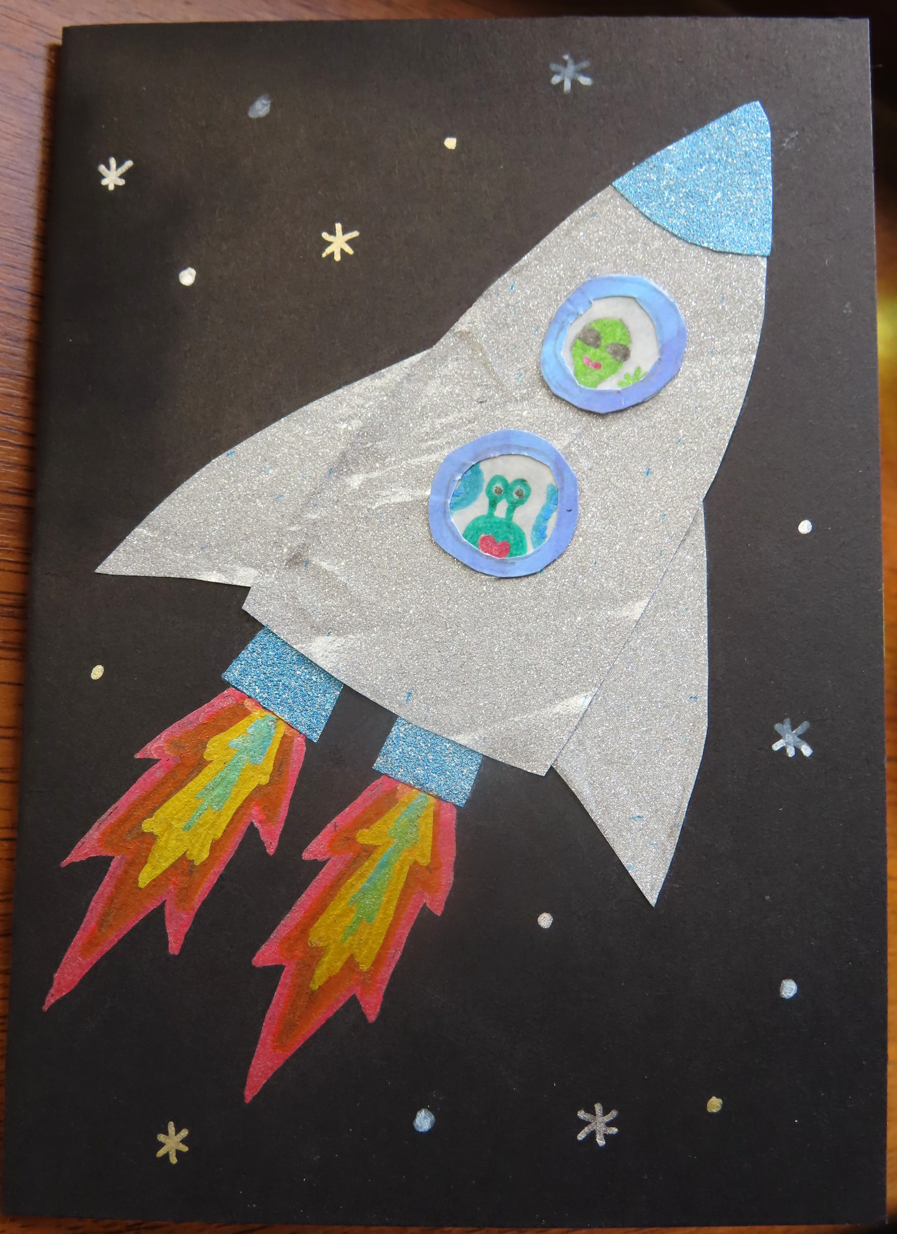 A handmade card depicting a silver rocket flying through space, with two green aliens in the windows waving