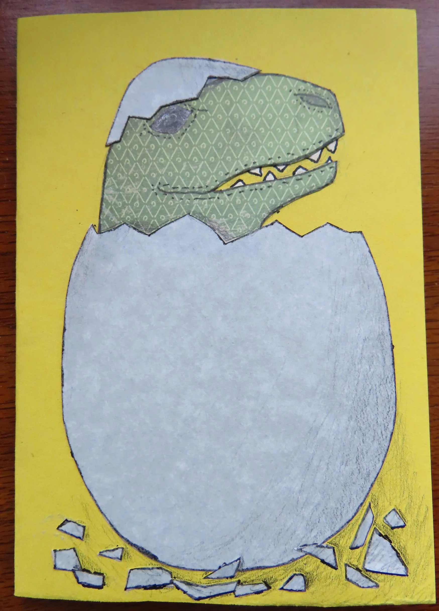 A handmade card depicting a baby T-Rex hatching from an egg on a yellow background