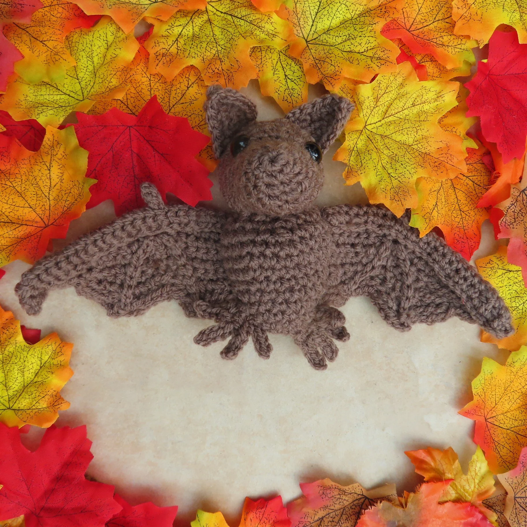 A crocheted bat with outstretched wings surrounded by autumn leaves in which the bat has made a 'leaf angel'