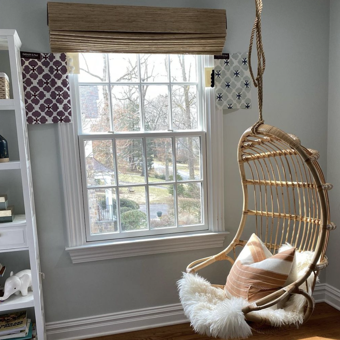 A cozy reading nook with a wicker hanging chair, a fluffy white throw, and decorative pillows next to a large window with patterned curtains. Outside, trees and a yard are visible.