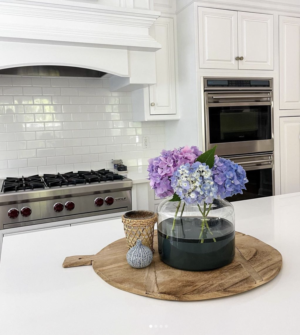 A kitchen with white cabinets and a white subway tile backsplash. In the foreground, a wooden cutting board on a white countertop holds a glass vase with purple and blue hydrangeas, a small woven cup, and a decorative small pear-shaped object.