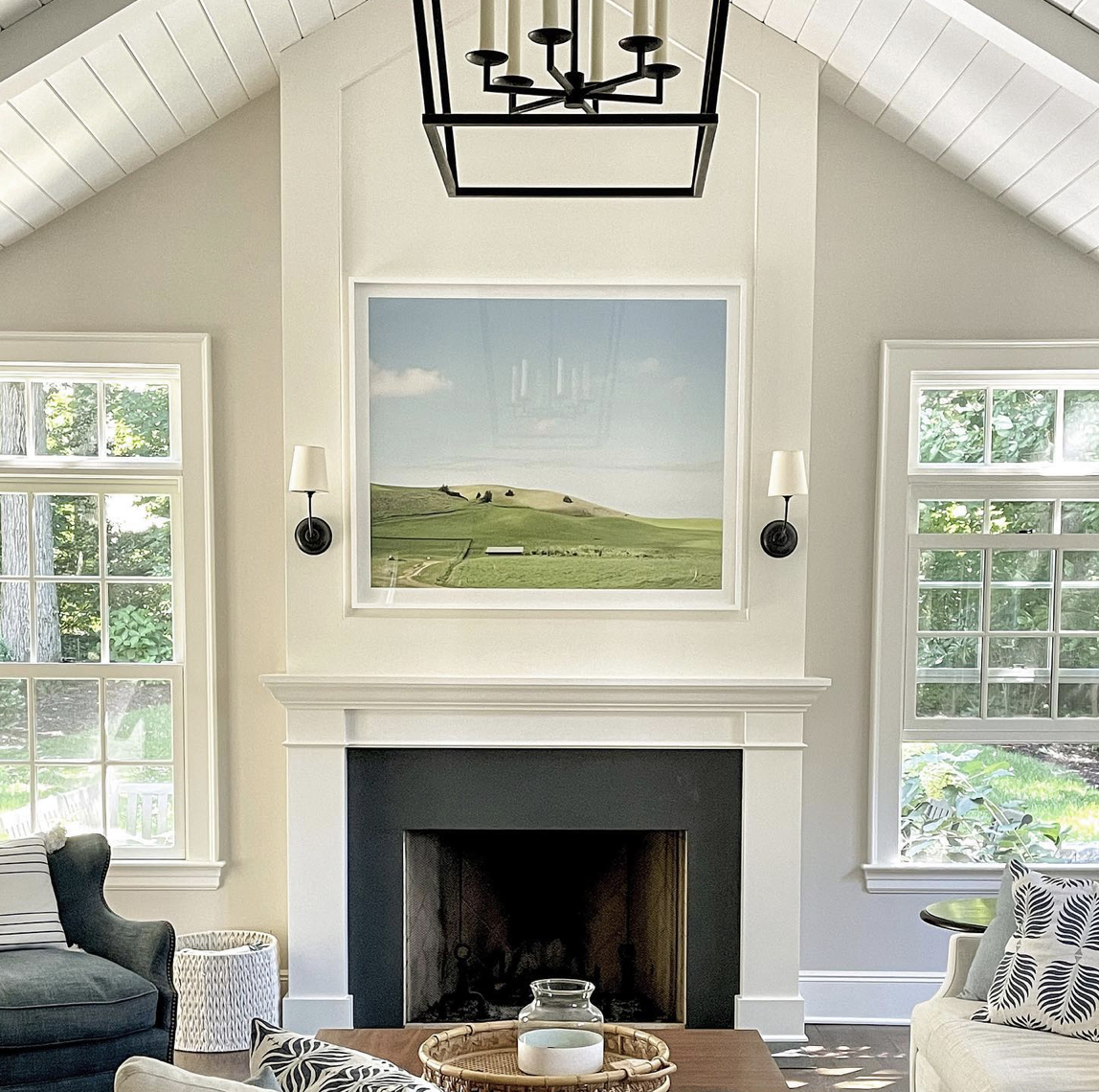 Living room with white walls, two large windows, a white fireplace with black accents, a large landscape painting above the fireplace, a chandelier hanging from the ceiling, and seating including a black armchair and a white armchair with patterned p