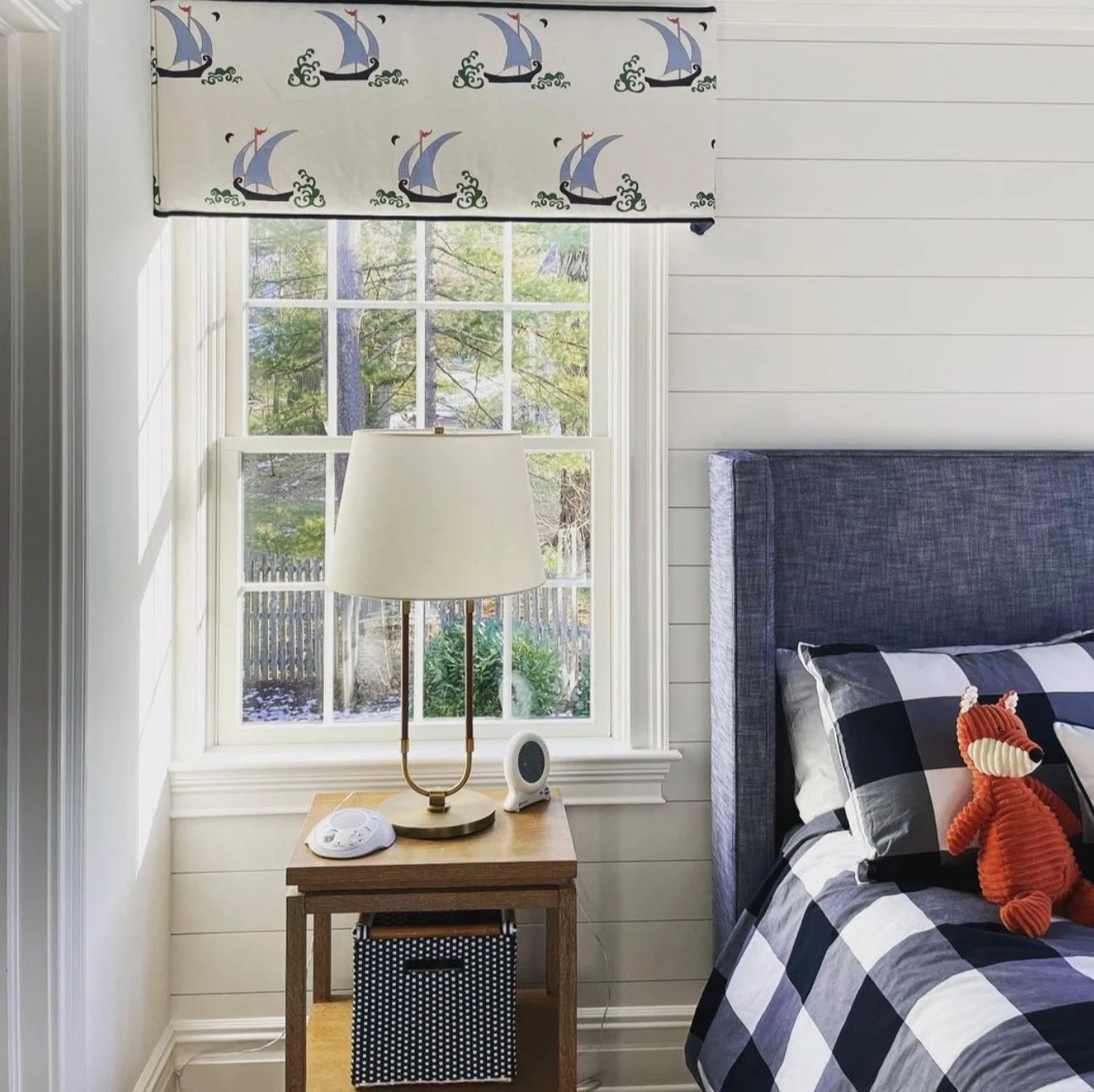 A bedroom corner with a window, a nightstand, a lamp, and a bed. The window has a sailboat patterned valance, the nightstand holds a white lamp, a small device, and a speaker. The bed has a checked blanket and a red plush toy.