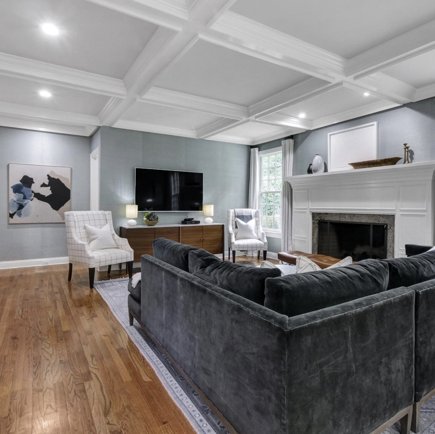 Living room with a dark gray velvet sofa, two white plaid armchairs, a wooden media console, flat-screen TV, fireplace with white mantel, artwork on the wall, and hardwood flooring.