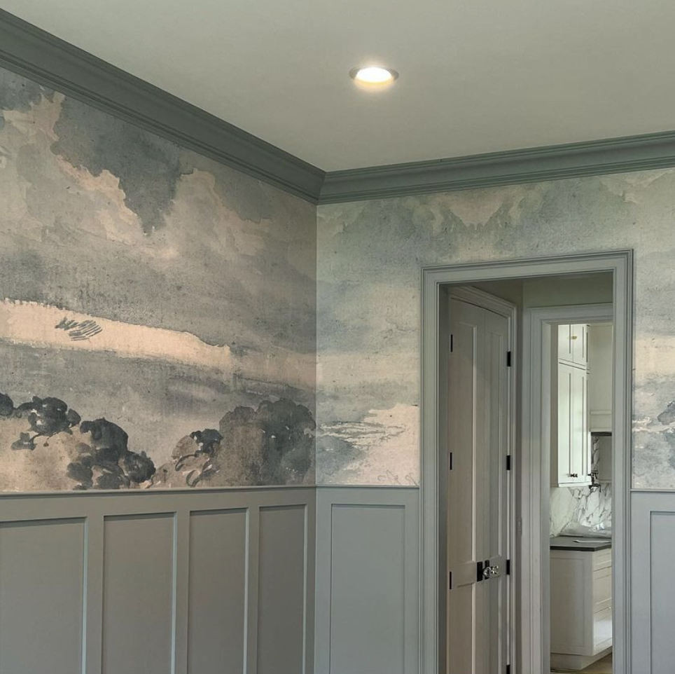 Interior view of a room with watercolor-style wall art, wainscoting, and a doorway leading to a kitchen with white cabinets and marble countertops.