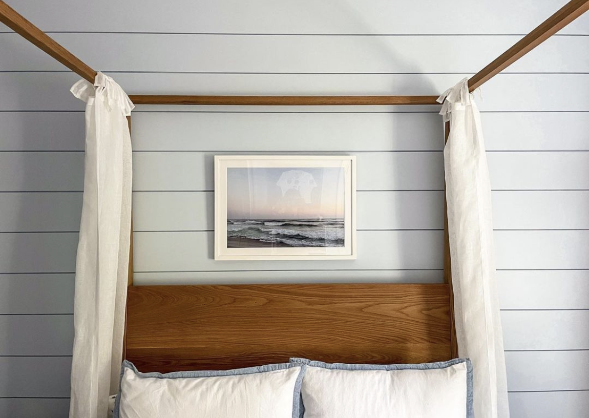A bedroom with light blue shiplap walls, a wooden headboard, and a framed picture of the ocean above the bed. Cream curtains hang from a canopy frame.