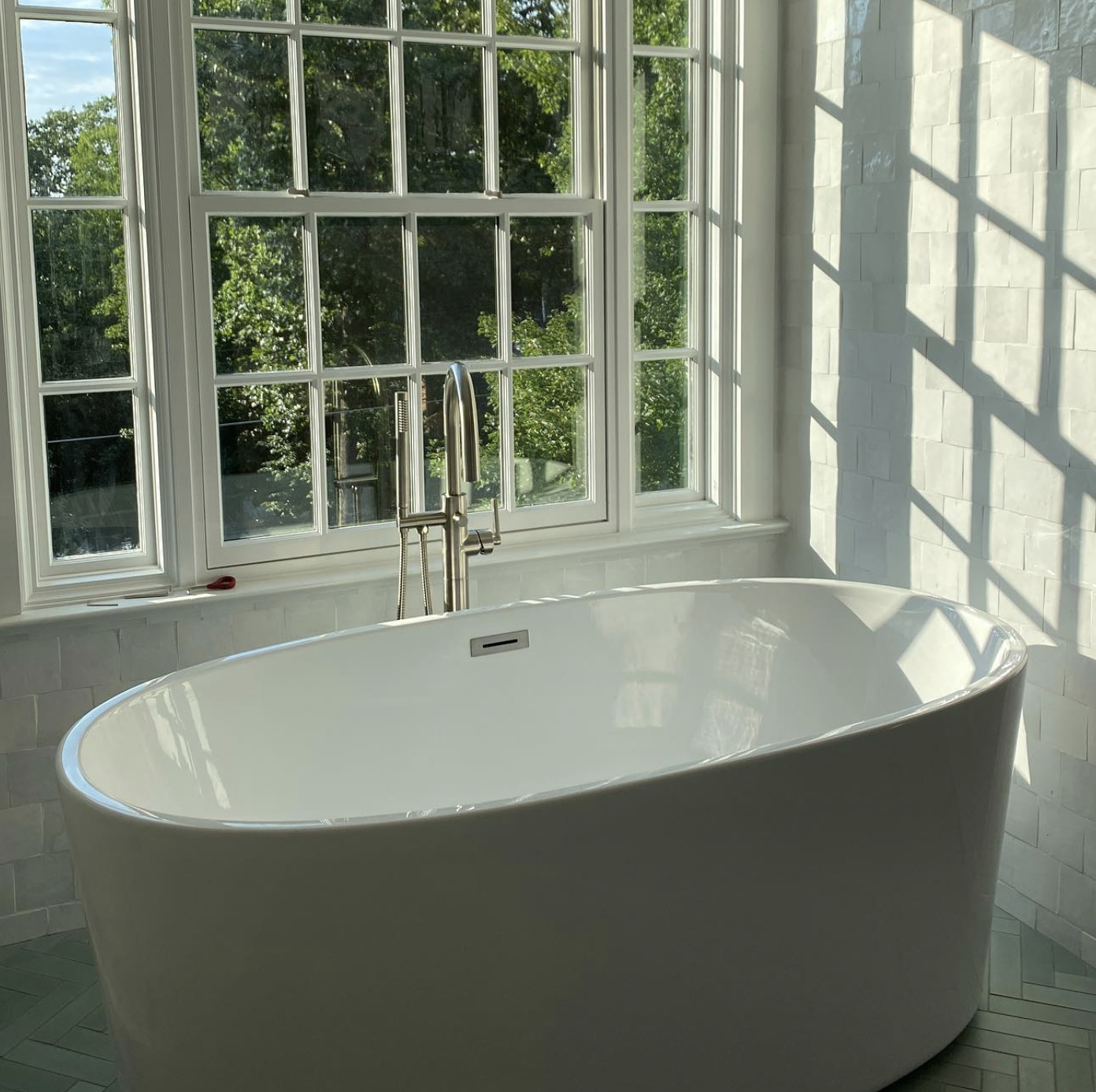 A modern bathroom with a white freestanding bathtub placed in front of large multi-pane windows, letting in sunlight and offering a view of green trees outside.