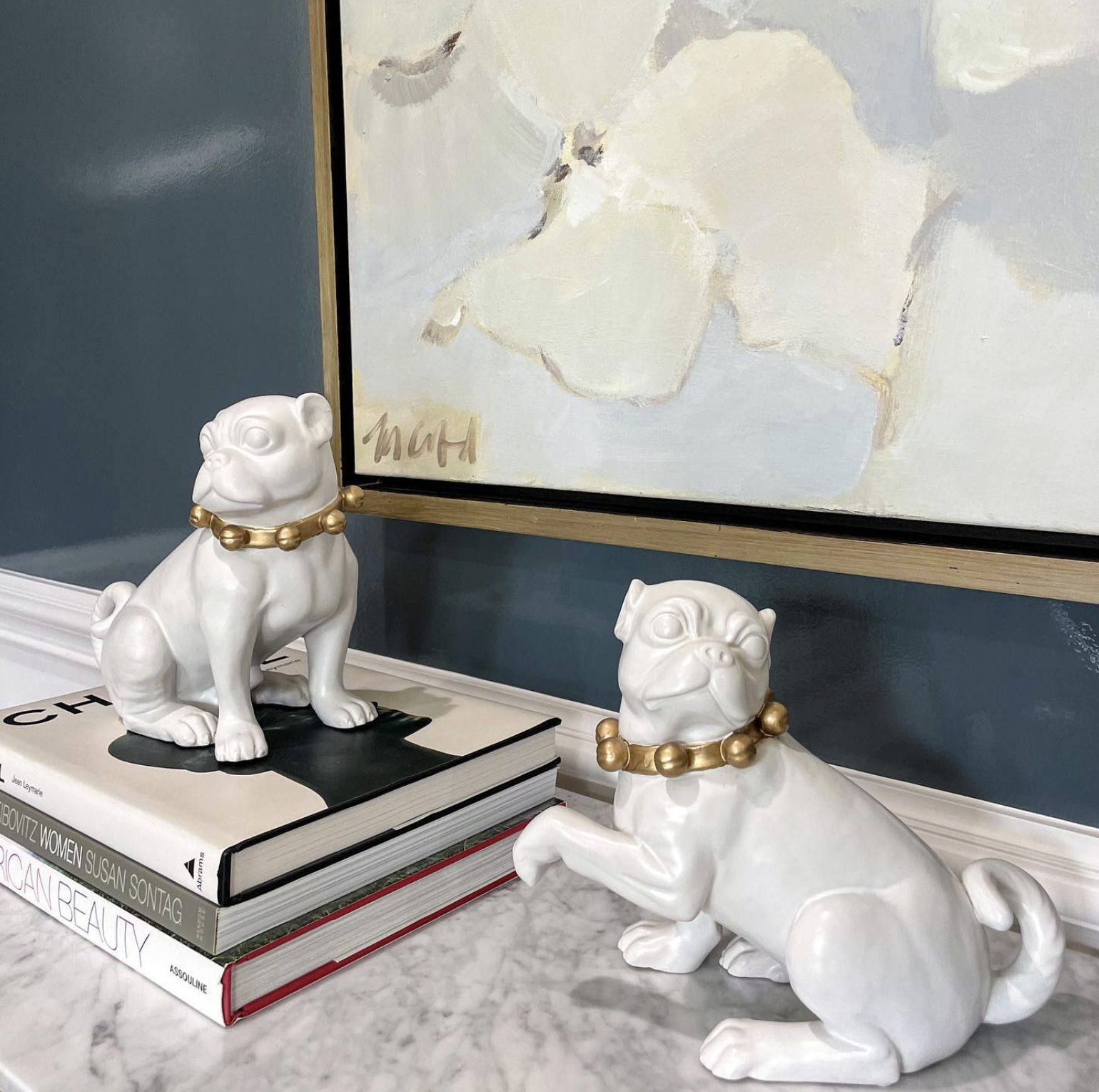 White ceramic dog statues with gold bead necklaces, one sitting on stacked books and the other standing on a marble surface, next to a painting of white flowers on a blue wall.