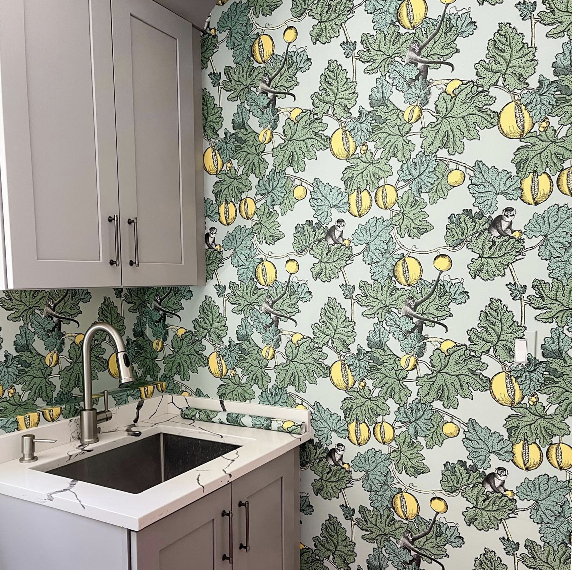 A kitchen corner with a grey cabinet, a marble countertop, a stainless steel faucet, and a sink. The wall is decorated with green leafy wallpaper featuring yellow pumpkins and small monkeys.