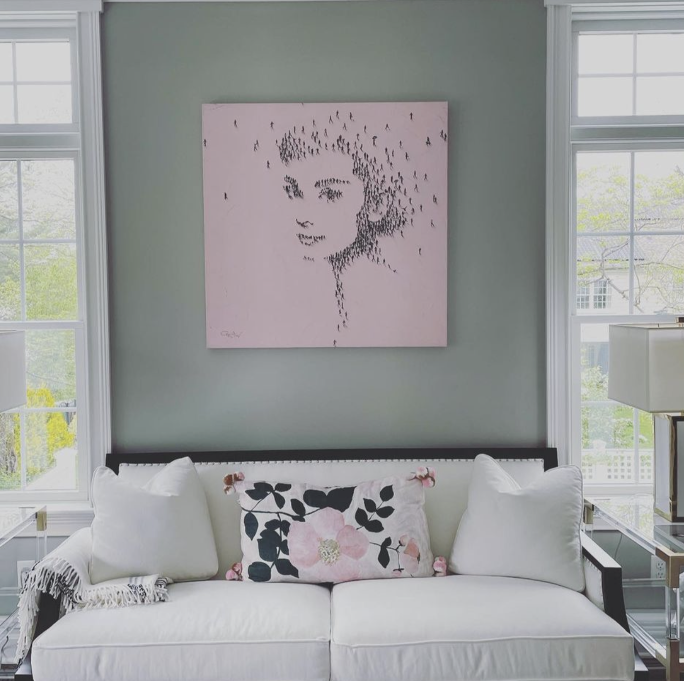 Living room with a white sofa, floral pillow, and wall art of a woman's face made with small dark figures.