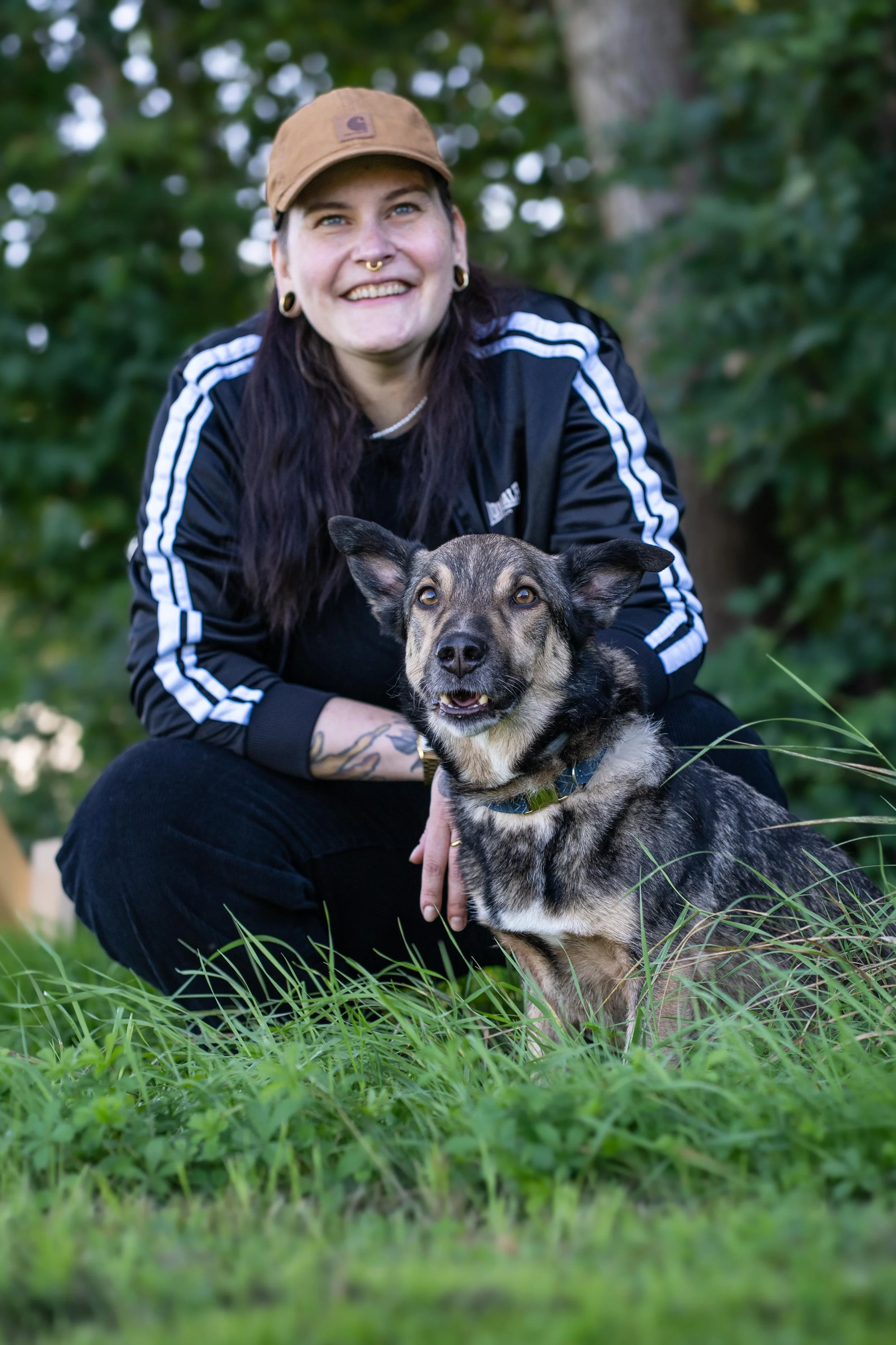 Junge Frau mit Tätowierungen und Ohrringen kniet im Grünen, trägt einen braunen Cap und lächelt, während sie einen Hund hält.
