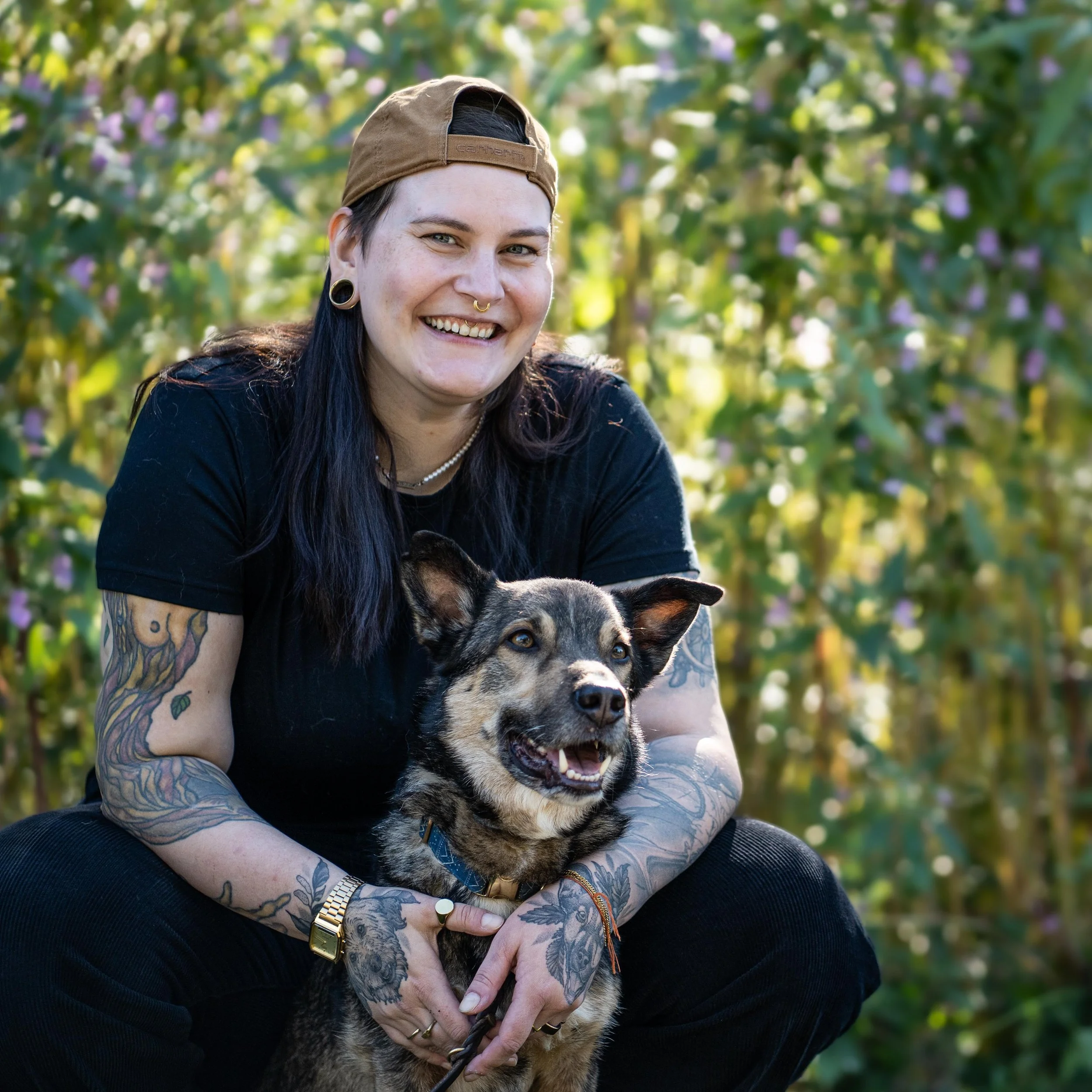 Junge Frau mit Tattoos und Piercings hält einen Hund in den Armen, draußen vor einem blühenden Garten.