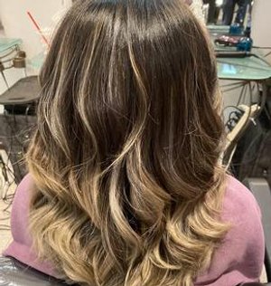 Woman with shoulder-length, wavy hair that is darker at the roots and gradually lightens to a blonde at the ends, seated in a salon.