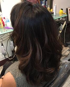 Back view of a woman with shoulder-length, wavy, dark brown hair with lighter highlights, sitting in a salon chair.