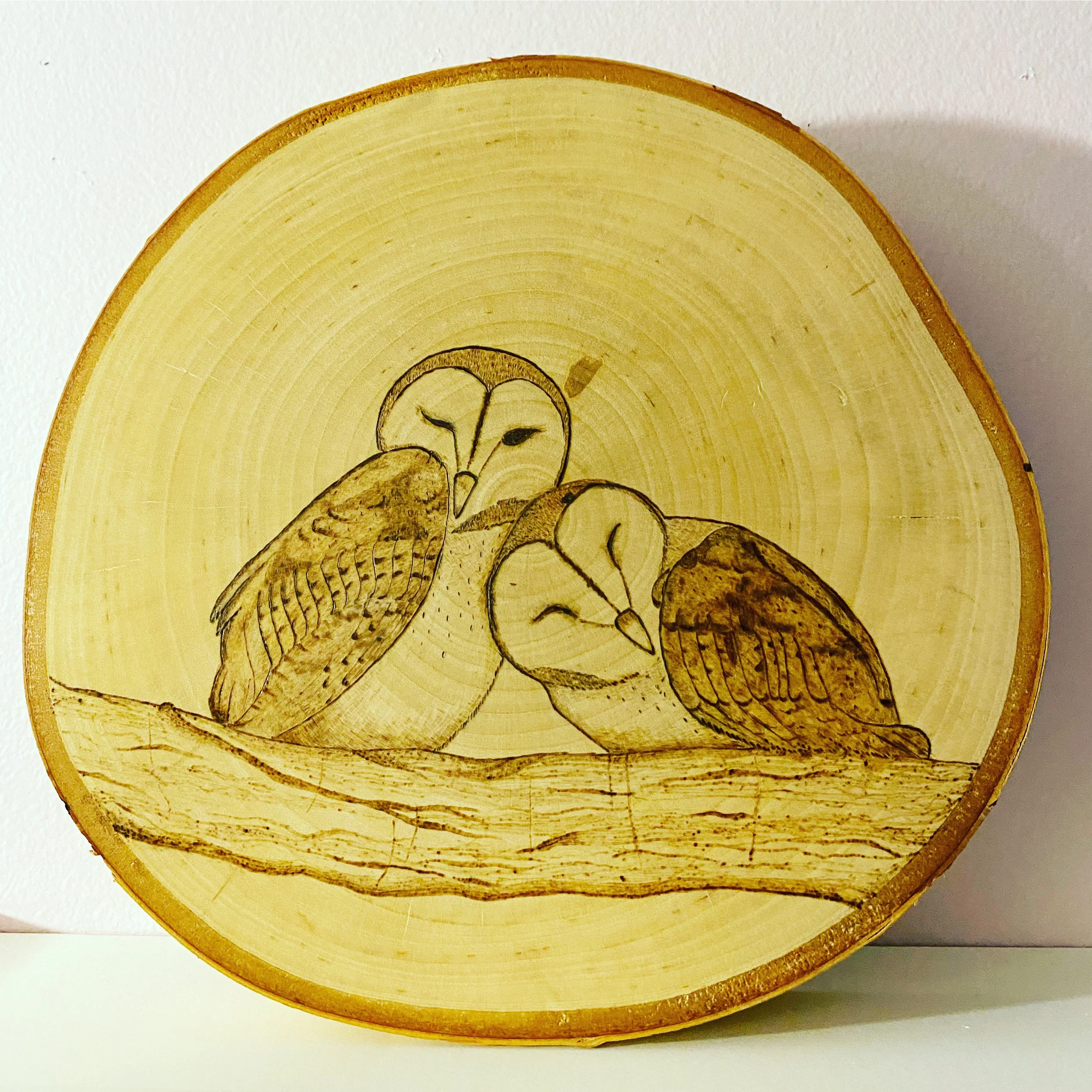 A round wood burned piece of two owls snuggling close on a tree branch.