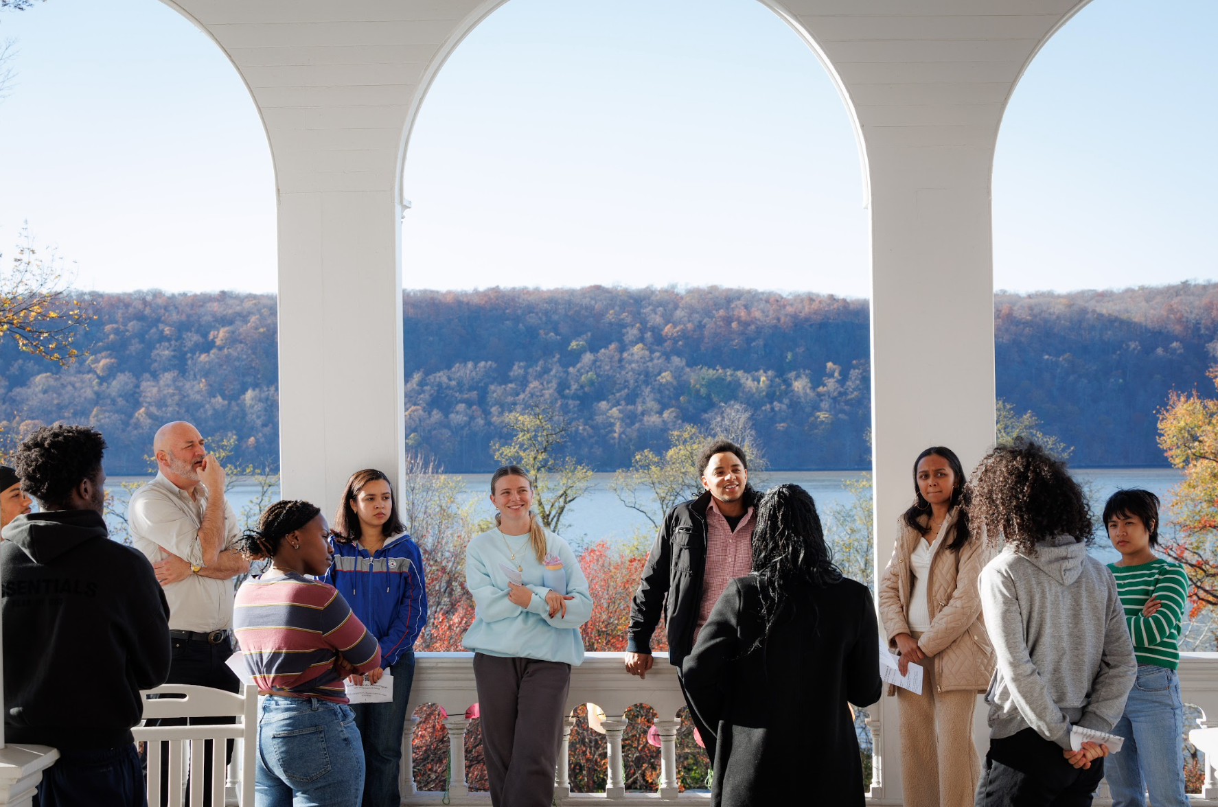 Group of people standing on a porch with an arched view of a river and wooded hills in the background.