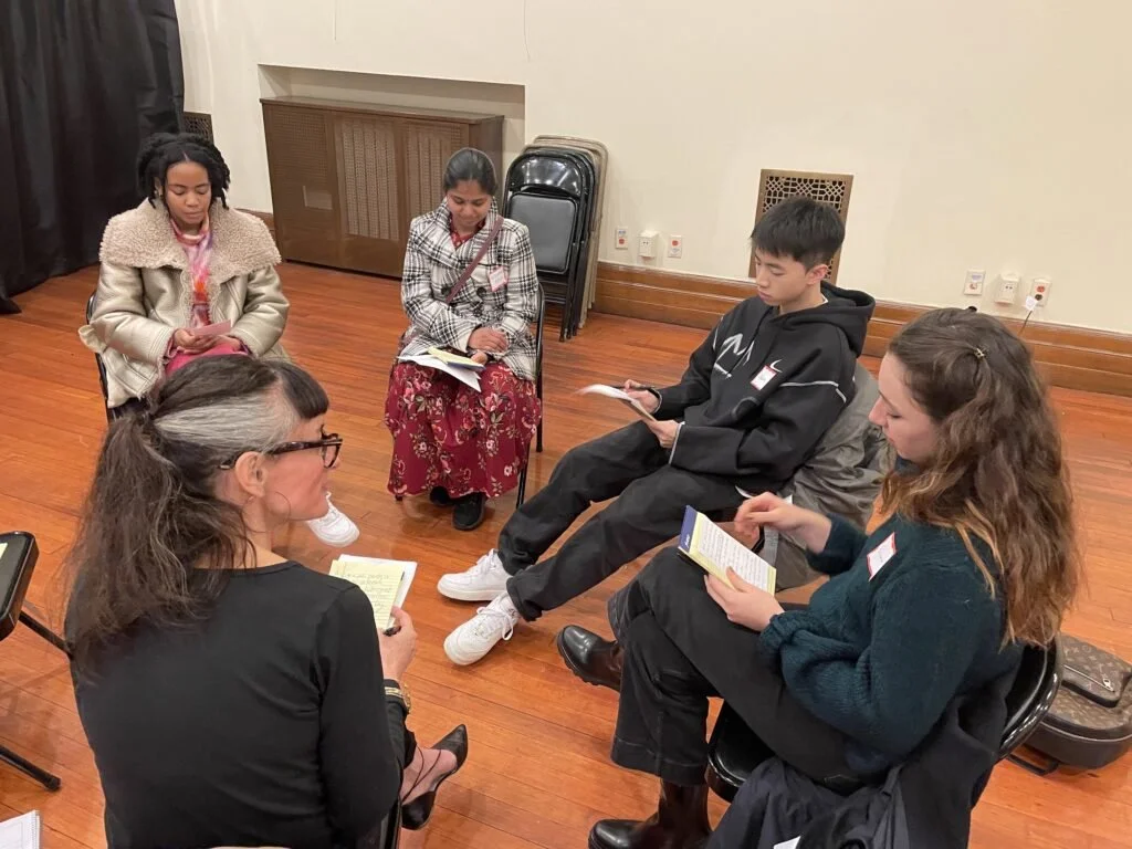 A group of five people sitting in a circle on chairs, engaged in a discussion or meeting. Some have notebooks or papers, suggesting a collaborative or educational setting. The room has a wooden floor and white walls with a stack of folded chairs in the background.