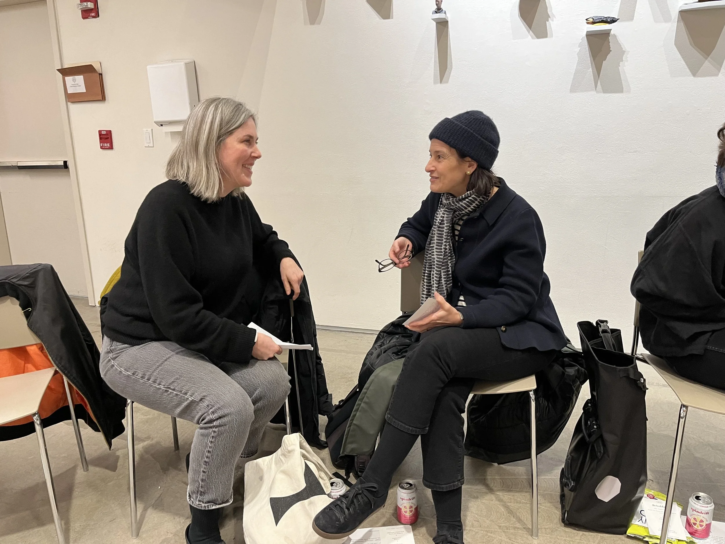 Two women seated and talking in a room, one with gray hair and the other wearing a beanie, surrounded by chairs and bags.