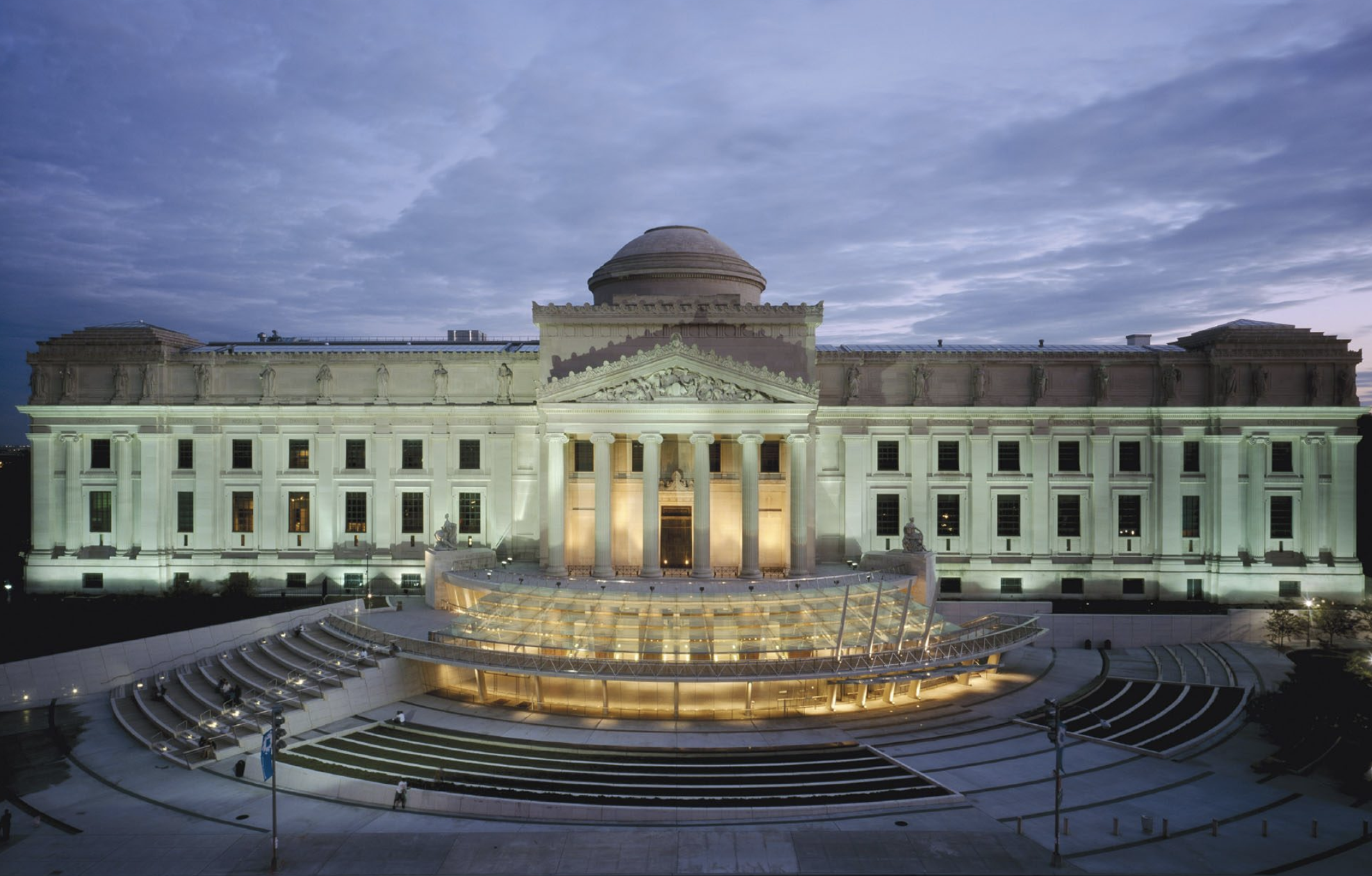 Exterior of a large, illuminated classical building with columns, dome, and steps leading to an entrance. The sky is dusk.