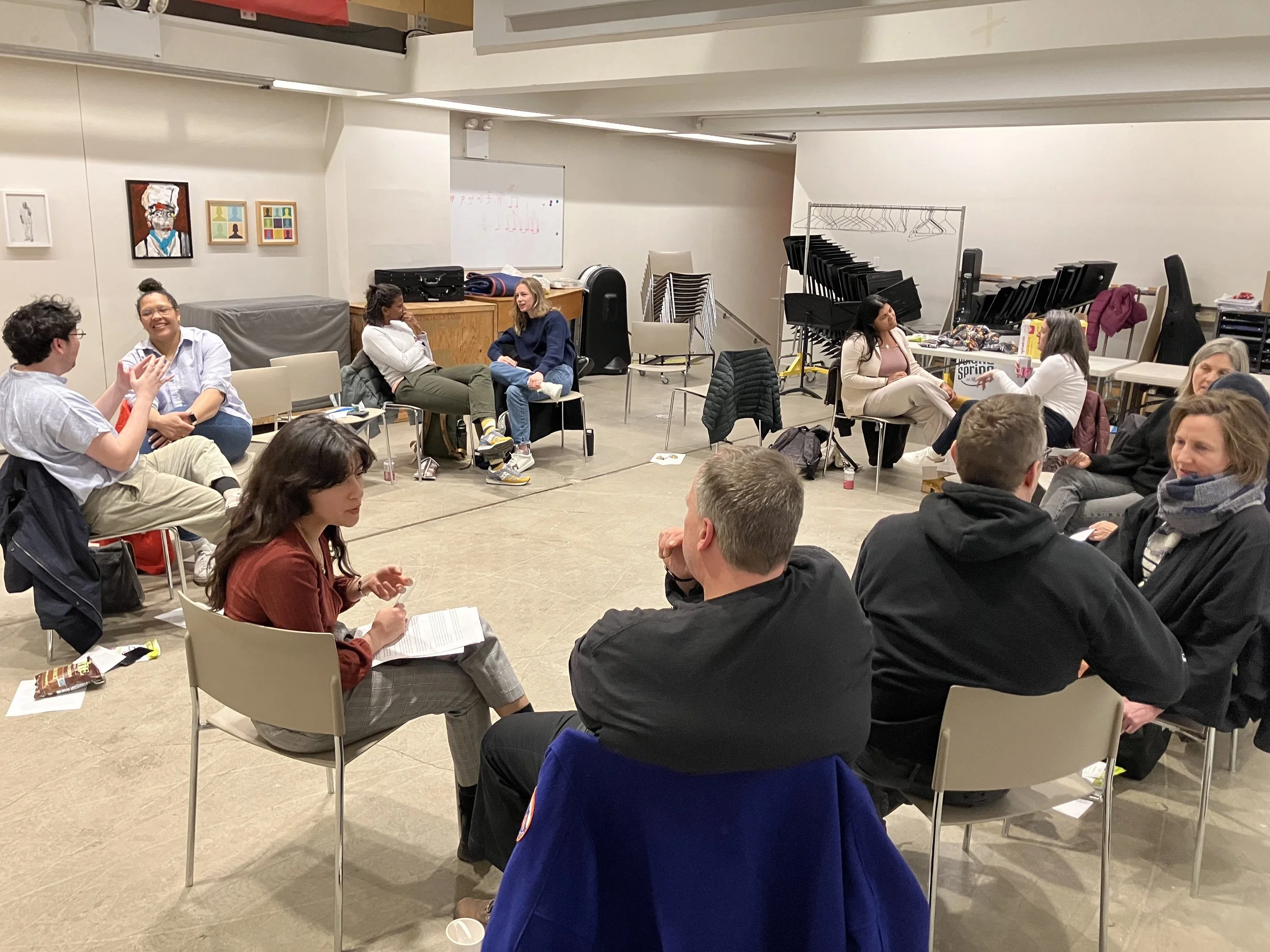 A group of people sitting on chairs in a circle in a room, engaging in a discussion or workshop. The setting appears to be casual and informal, with papers and refreshments on side tables. The participants are engaged in conversation, sitting in small groups.