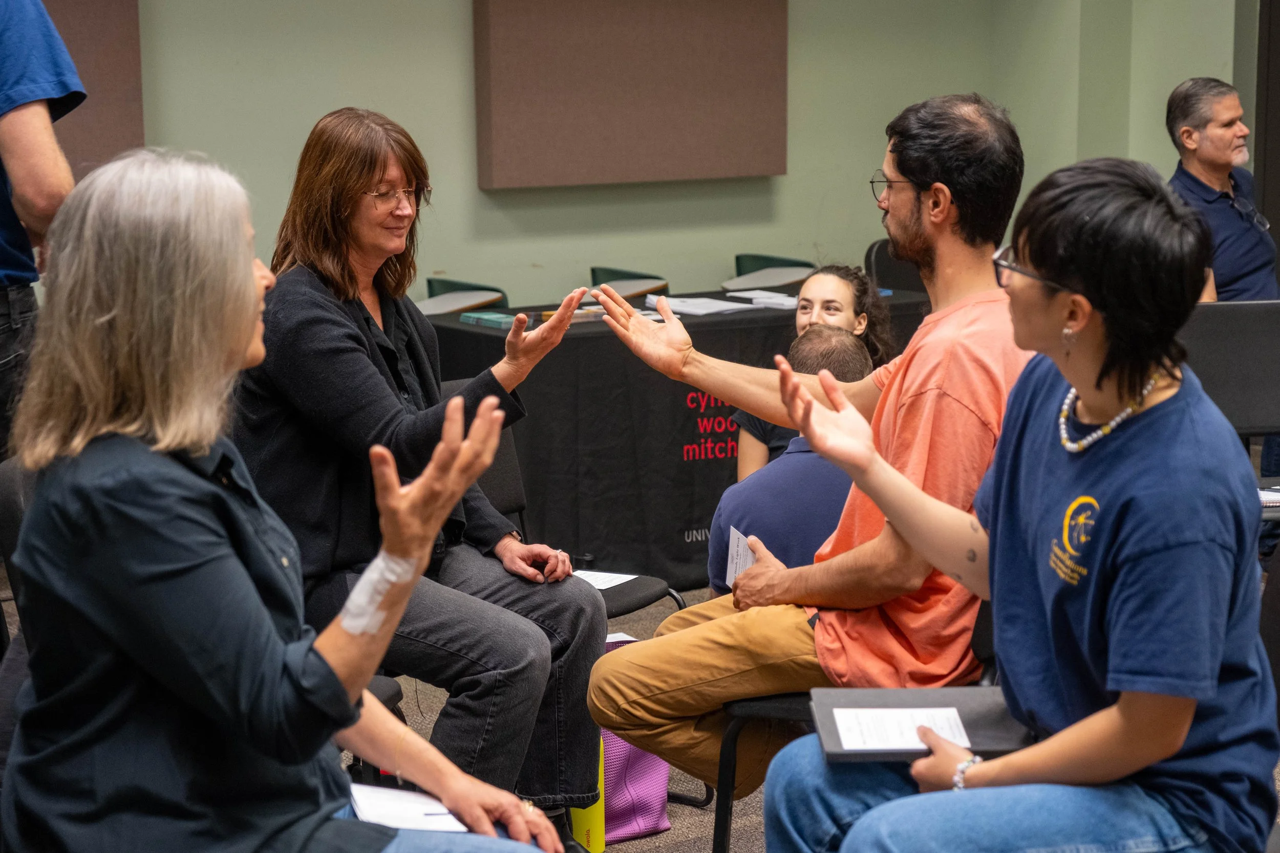 Group of people participating in a mindfulness or communication exercise, sitting in chairs, extending their hands towards each other in a classroom setting.