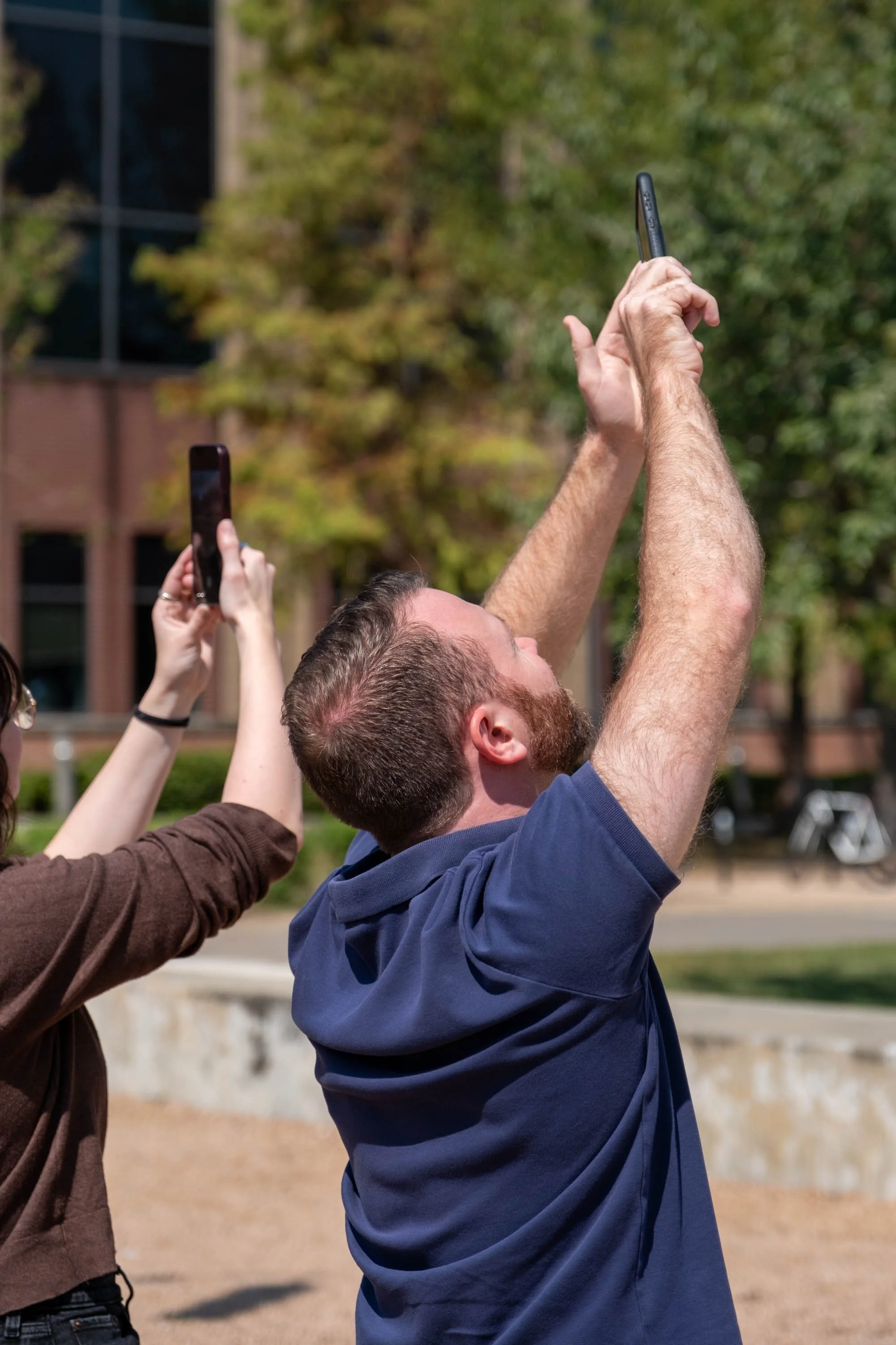Two people taking photos upwards with smartphones outdoors.