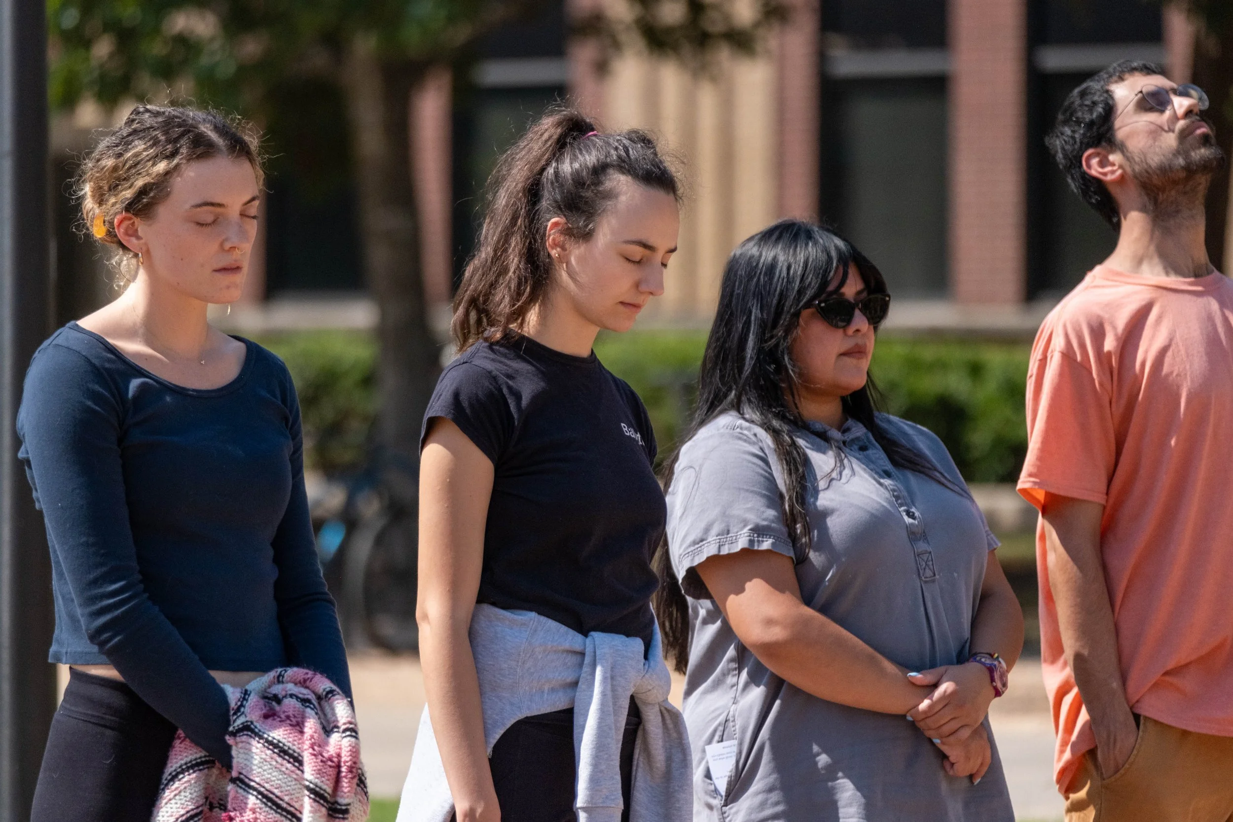 Four people standing with eyes closed outdoors, appearing contemplative, with trees and a building in the background.
