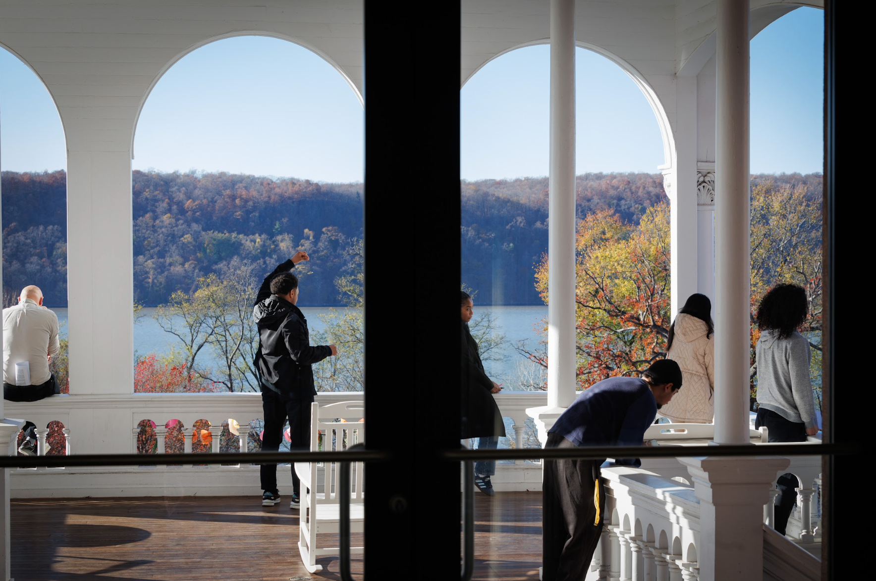 People on a veranda overlooking a lake and autumn trees through white arches.