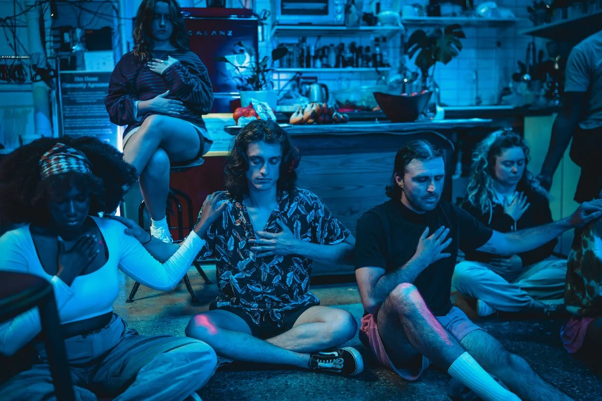 A group of people sitting cross-legged on the floor with eyes closed, participating in a meditation or mindfulness session in a dimly lit room with a blue hue.