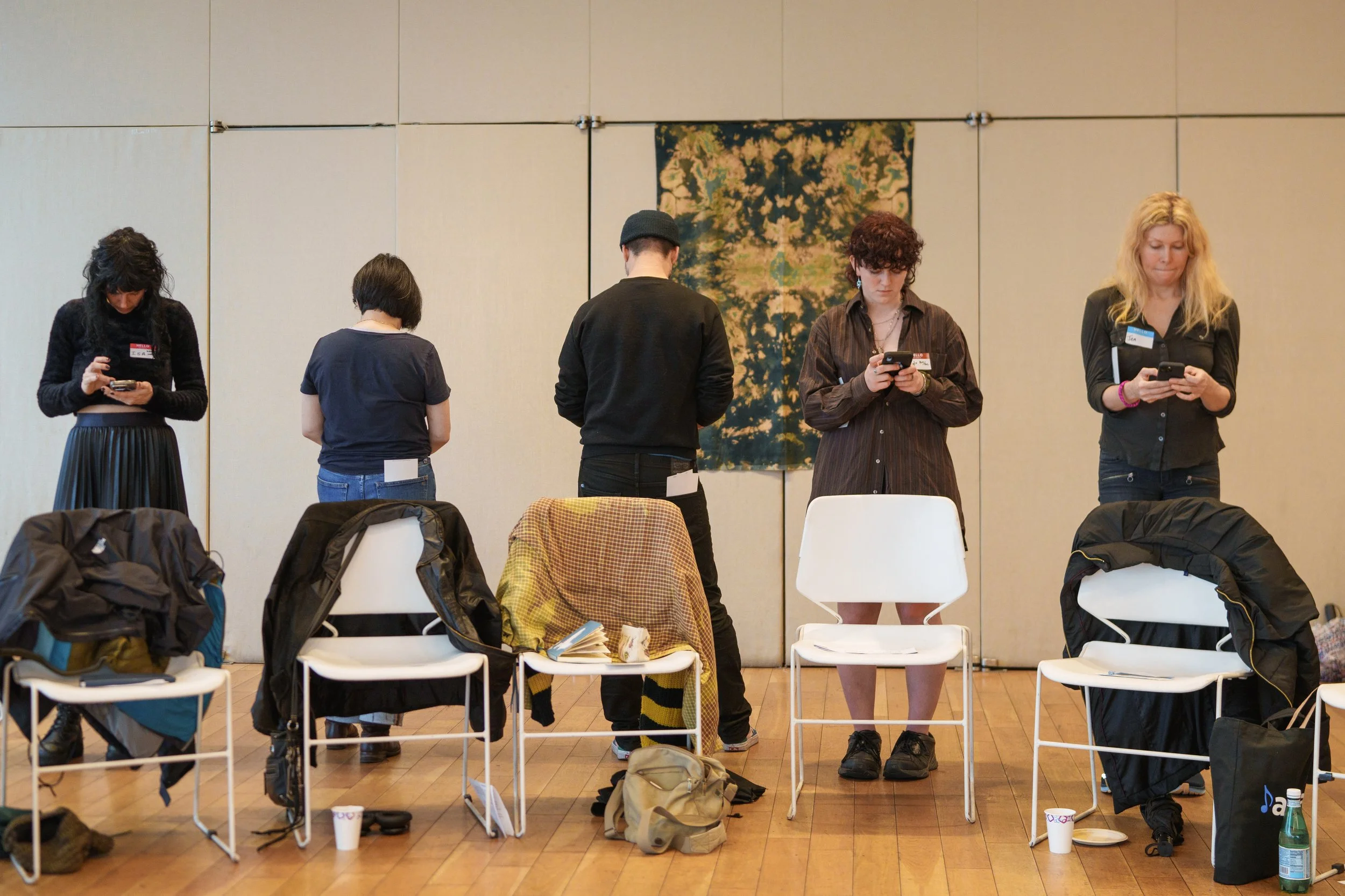 Five people standing behind chairs, using smartphones, in a room with a wood floor.