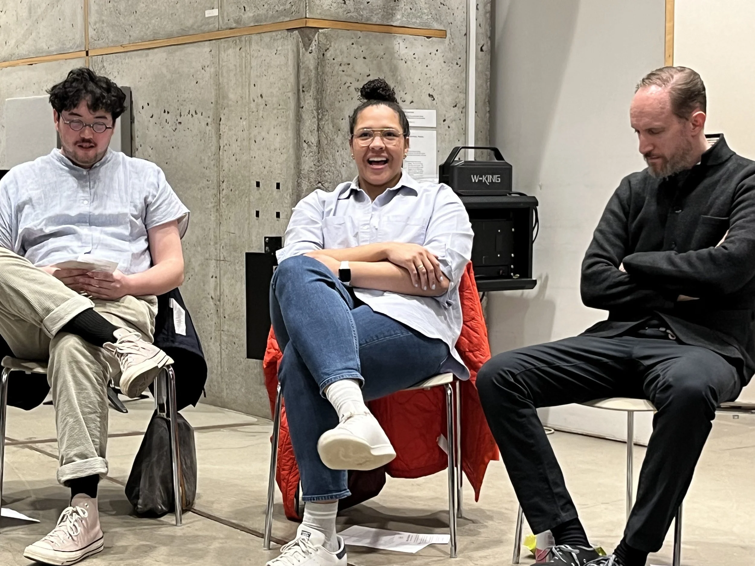 Three people sitting on chairs in a casual setting, with two of them smiling and engaged in a conversation, while the third looks down. They are in a room with concrete walls and a speaker in the background.