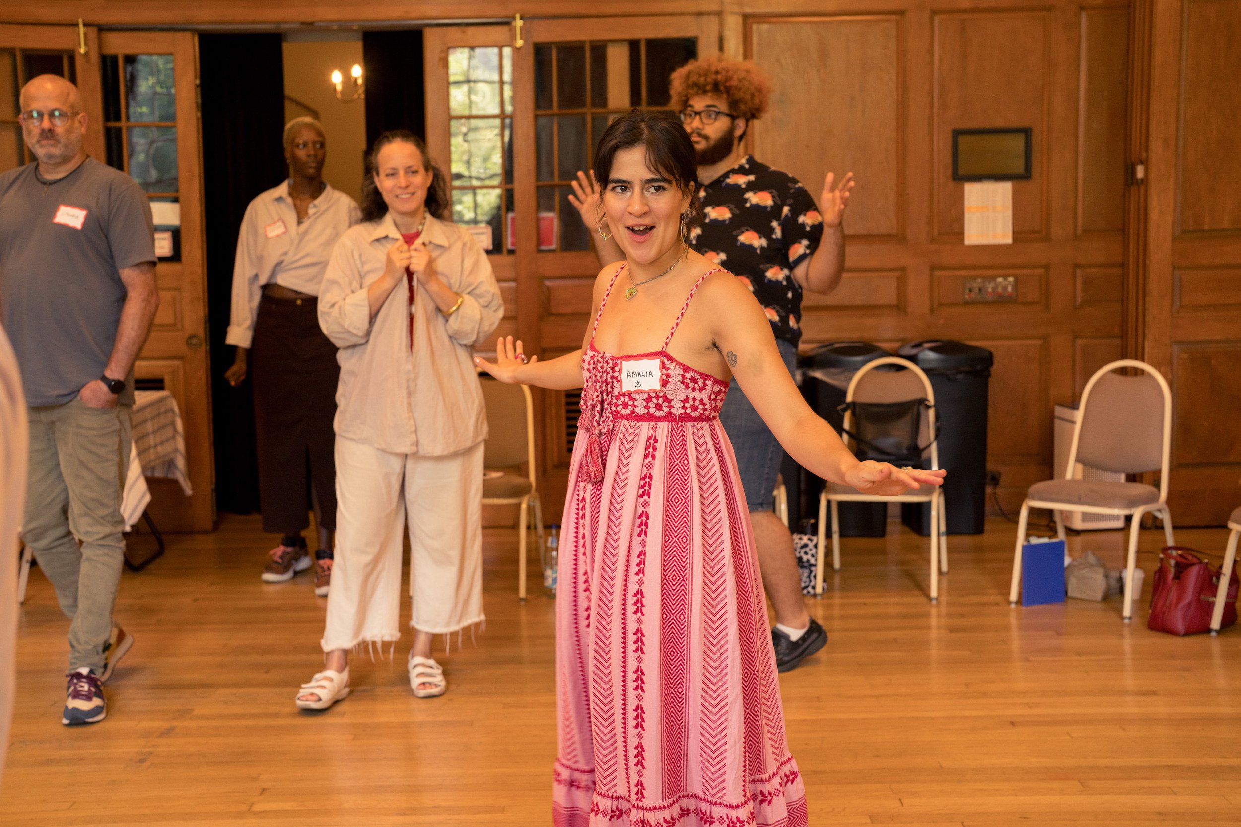 A group of five people standing in a room with wooden walls and floors, some smiling and wearing name tags. One person in the foreground is wearing a red and white patterned dress, with arms outstretched, seemingly talking or performing. Others are casually dressed, standing in a semi-circle, participating or observing.