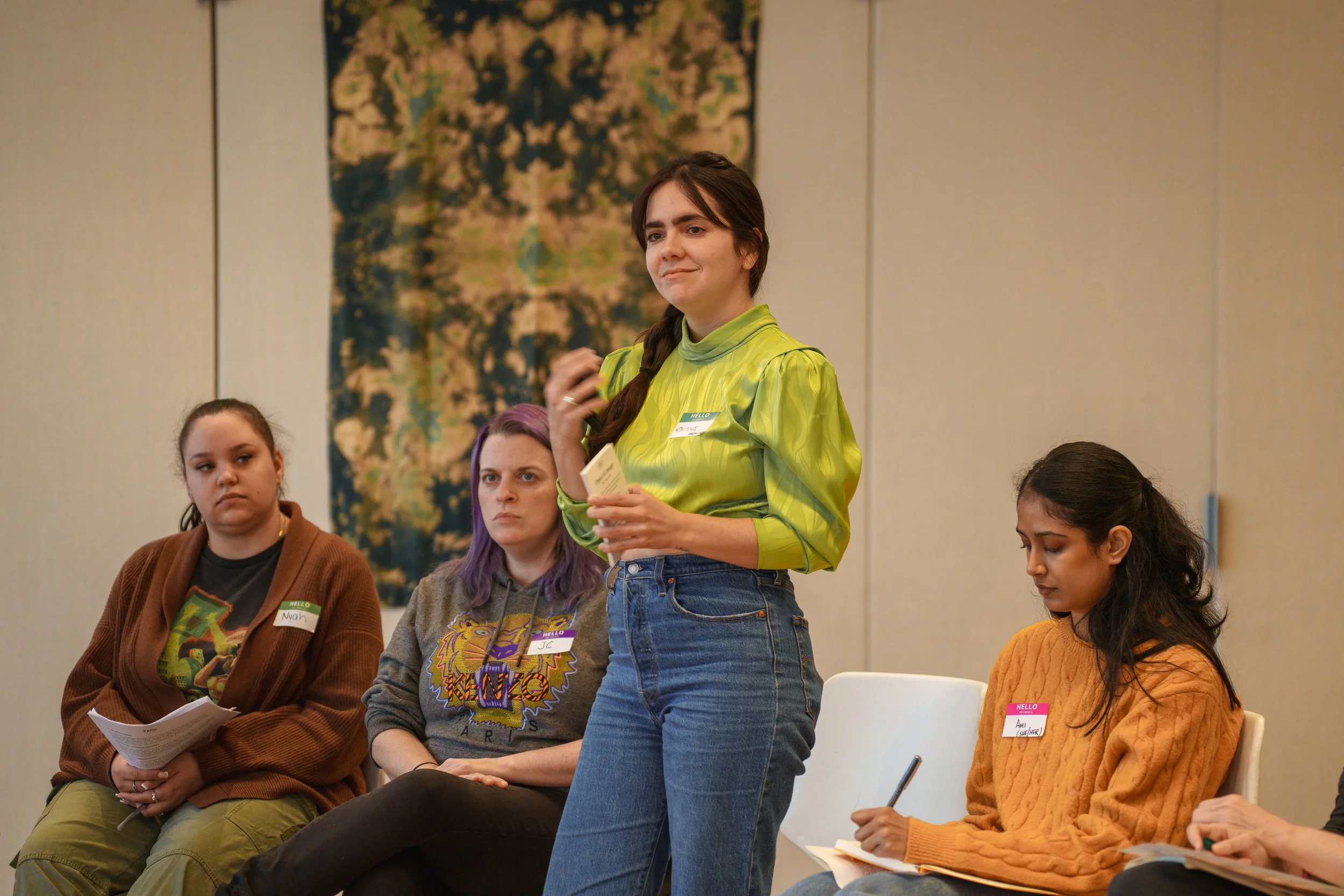 A group of people in a discussion session. A person in a bright green blouse is standing and speaking, while others are seated, listening, and taking notes. Name tags are visible, and they are indoors with a decorative tapestry in the background.
