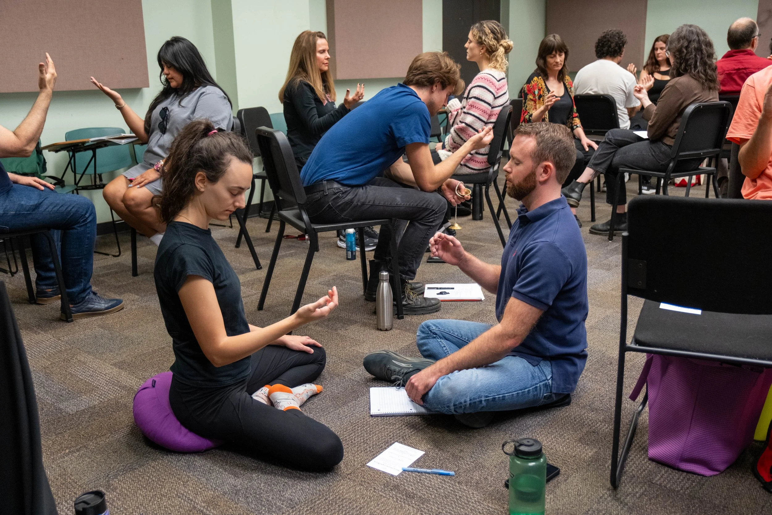 A group of people sitting in pairs engaging in a meditation or mindfulness exercise in a classroom setting.