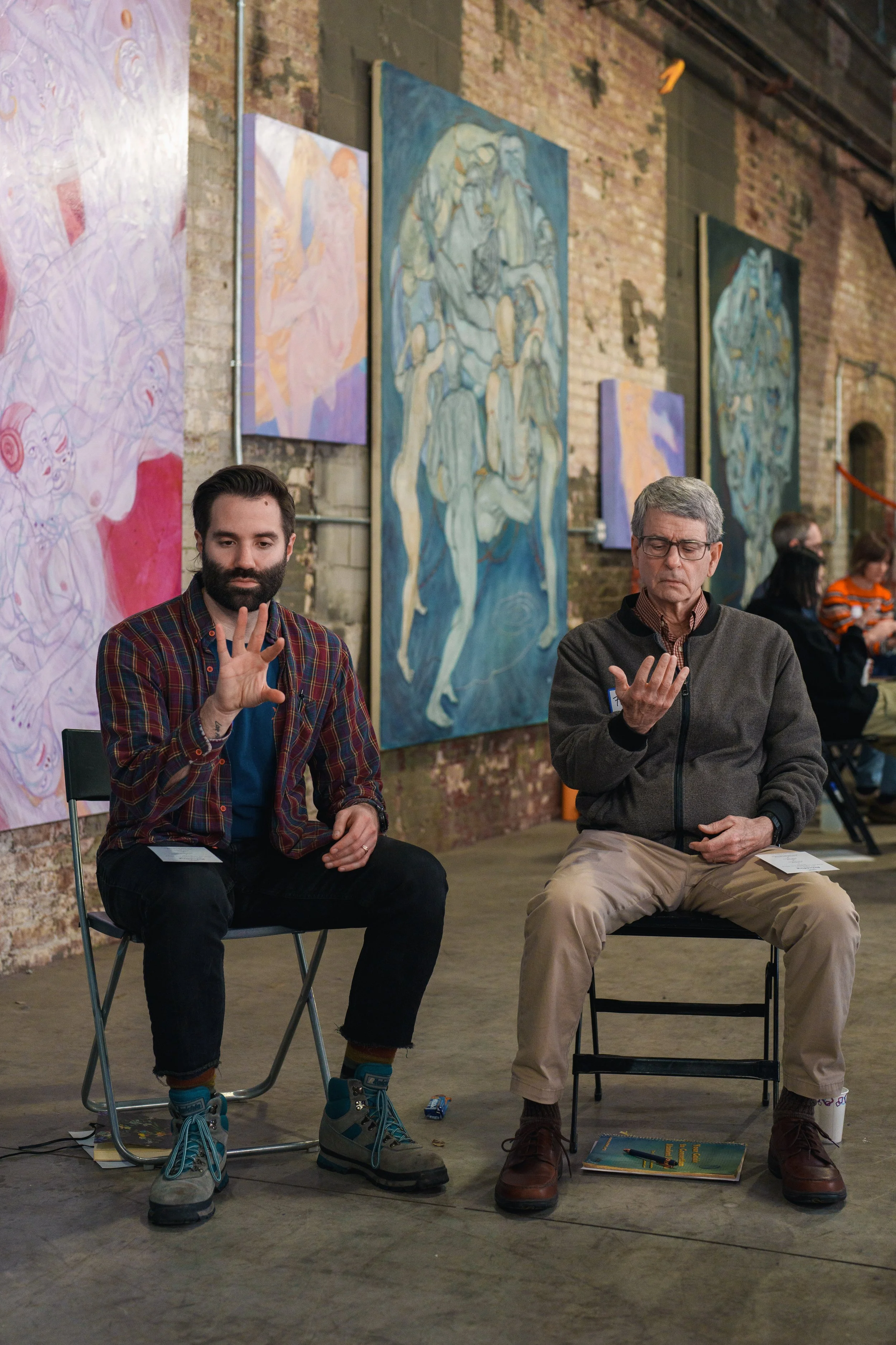 Two men sitting on chairs in an art gallery, displaying hand gestures, with abstract paintings on the walls behind them.