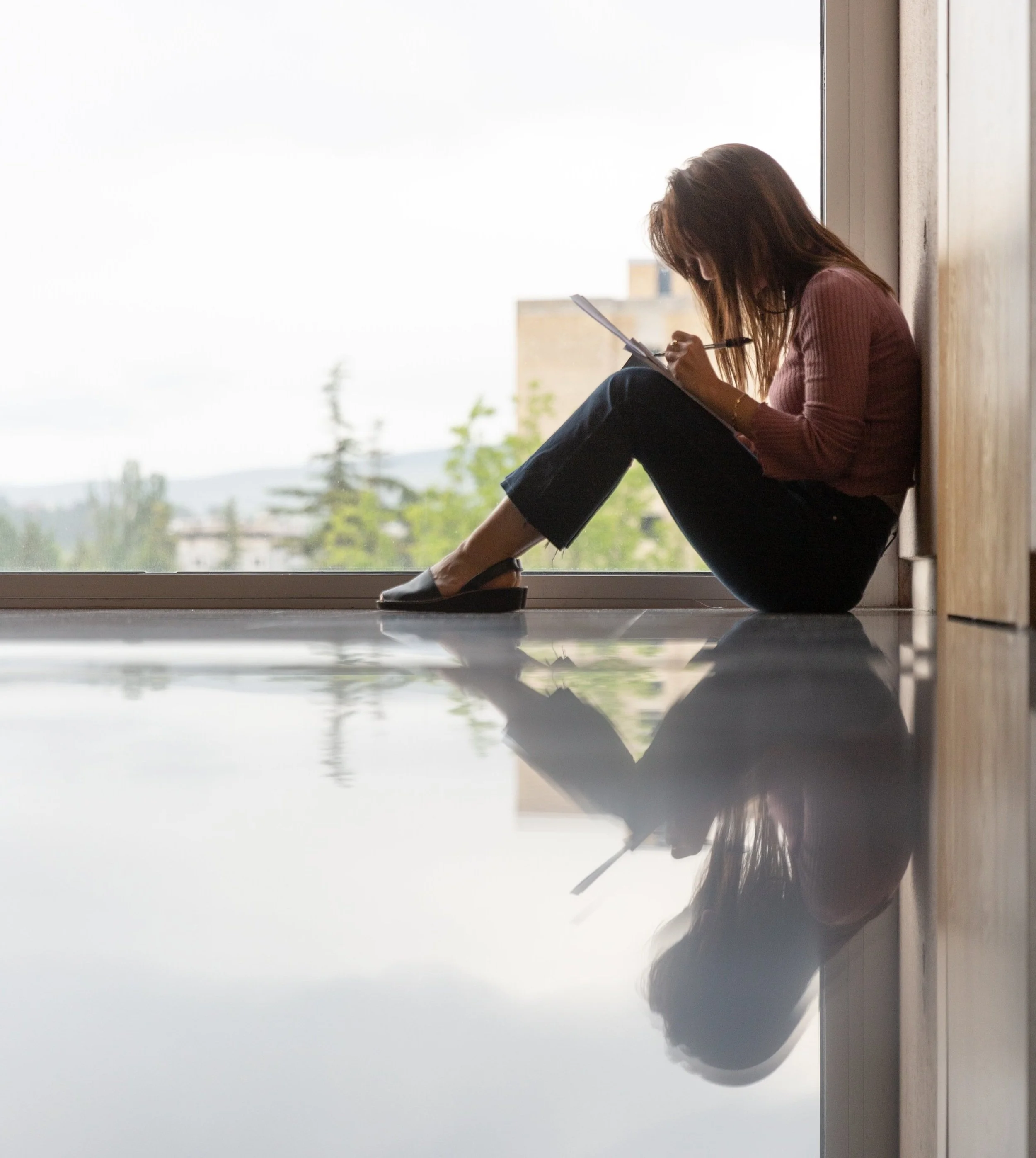 Woman sitting on the floor by a window, writing in a notebook, with her reflection on the polished floor.