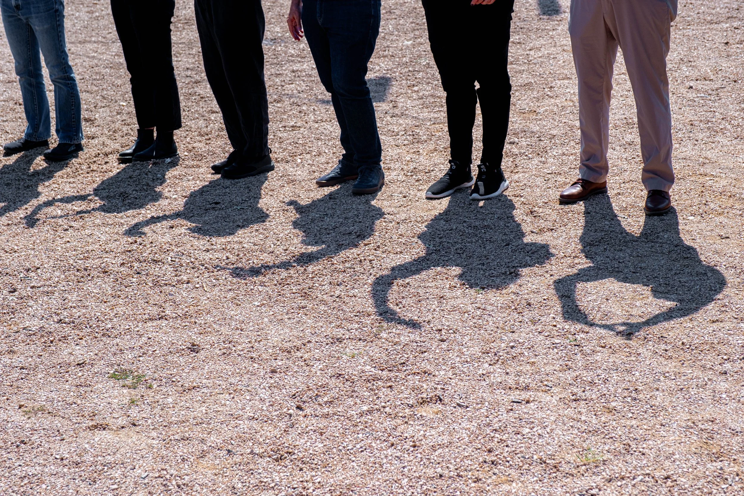 Legs of people standing on gravel with distinct shadows forming different hand shapes on the ground.