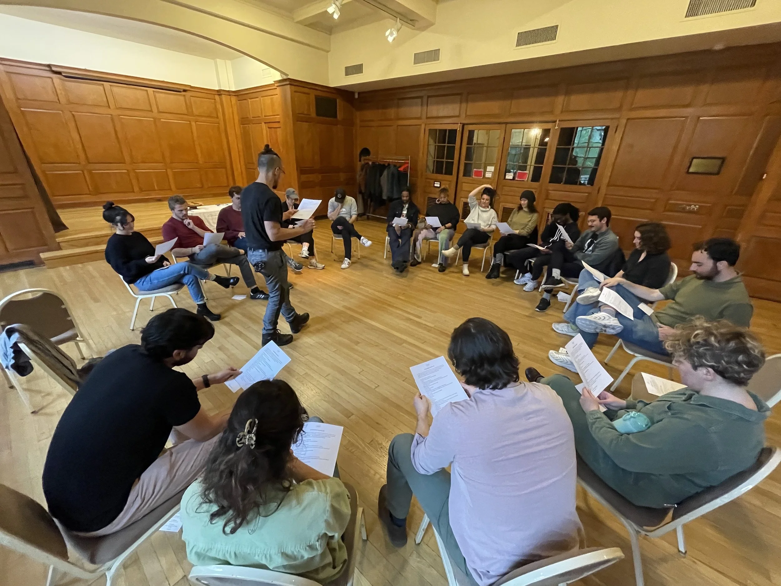 A group of people sitting in a circle in a room with wooden paneling, participating in a reading or workshop session, holding papers.