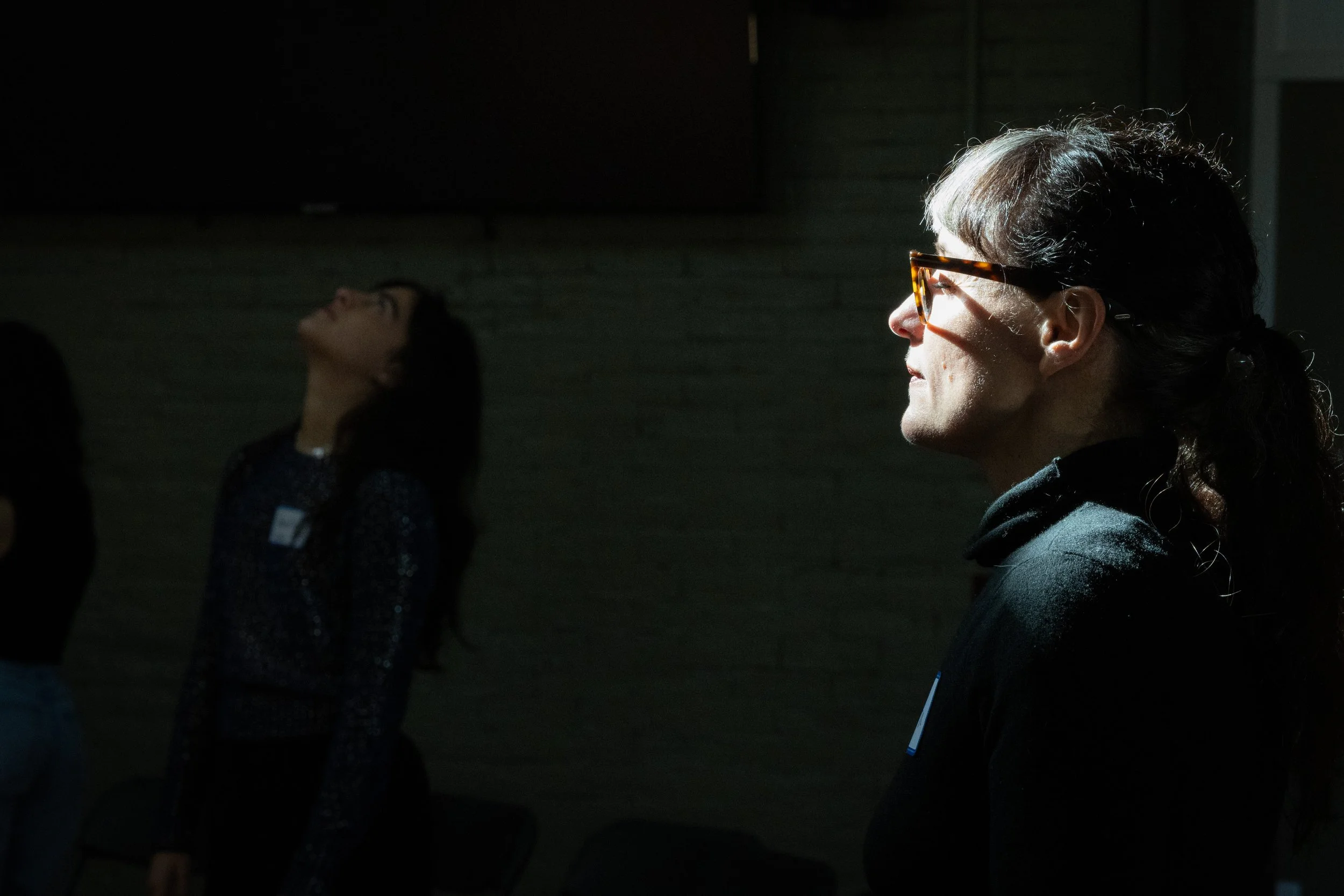 People standing indoors, one person in profile wearing glasses, another looking upward.