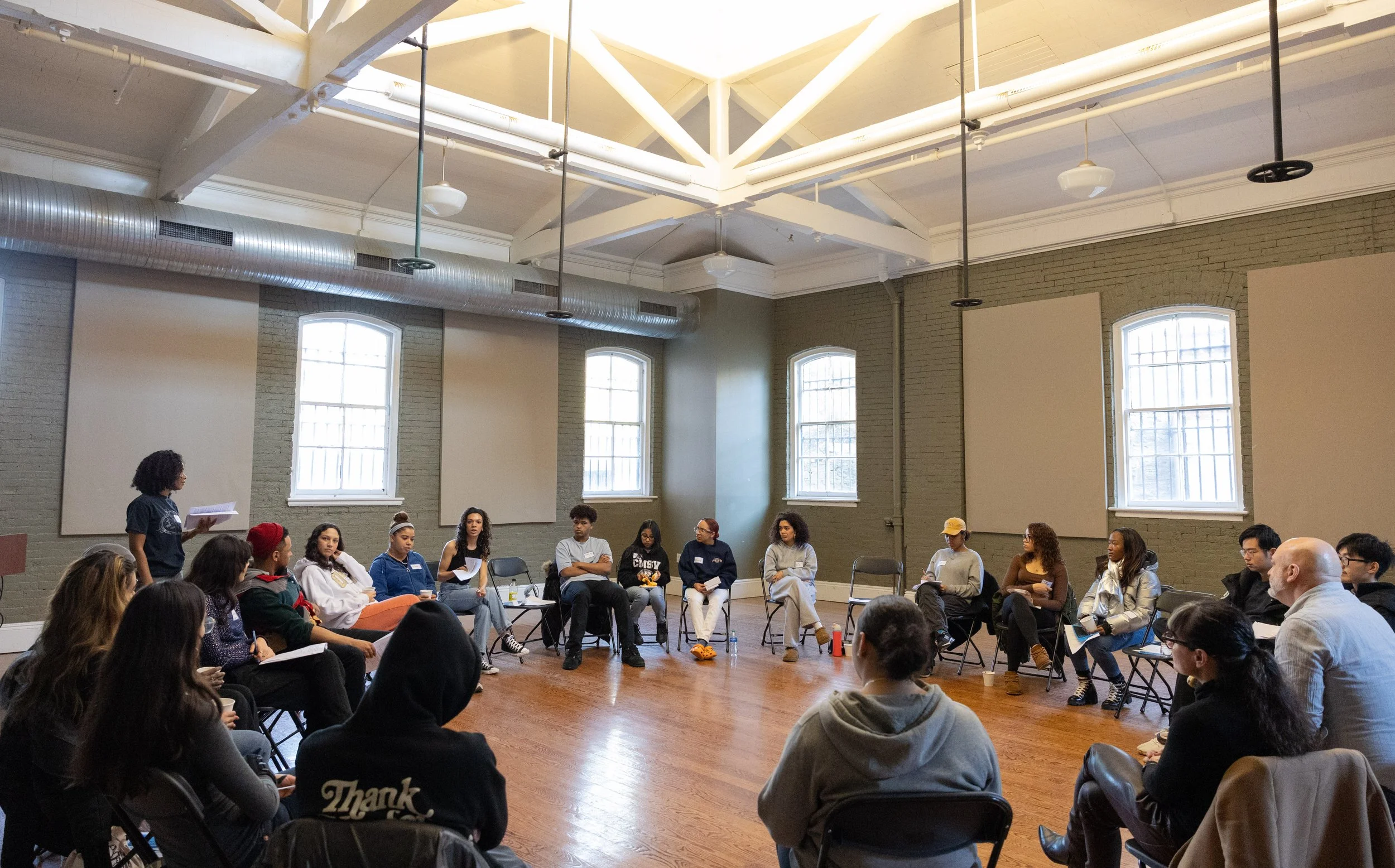 A group of people sitting in a circle in a large room with high ceilings and exposed beams. The attendees are engaged in a discussion, with one person standing to speak while others sit, some taking notes. The room has windows with natural light and the floor is hardwood.