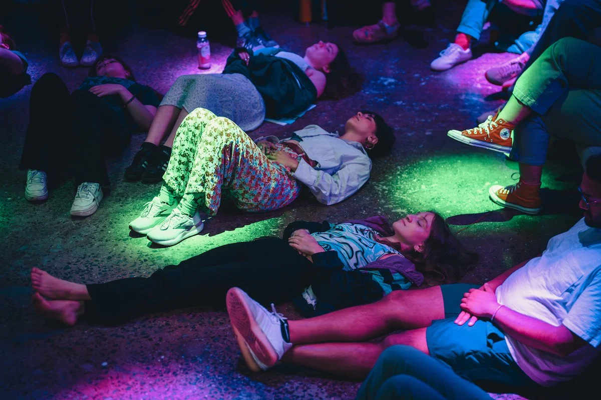 People lying on the floor under colorful lights, appearing relaxed, possibly in a group meditation or relaxation setting.