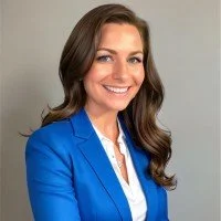 A woman with long brown hair wearing a blue blazer and white blouse, smiling for a professional portrait.