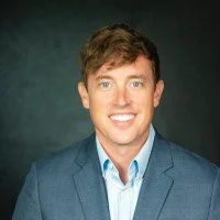A young man with brown hair and blue eyes, wearing a blue blazer and light blue shirt, smiling against a dark background.