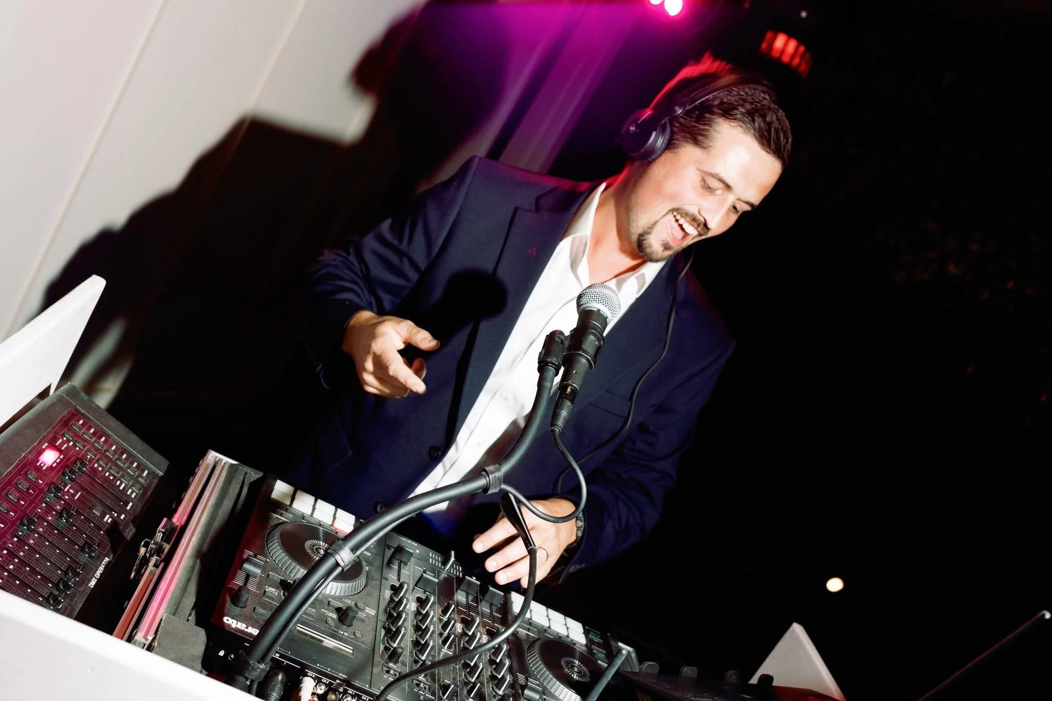 A DJ in a white shirt with rolled-up sleeves, wearing headphones, standing behind a DJ setup with a laptop, at an indoor event with stage lighting and speakers.