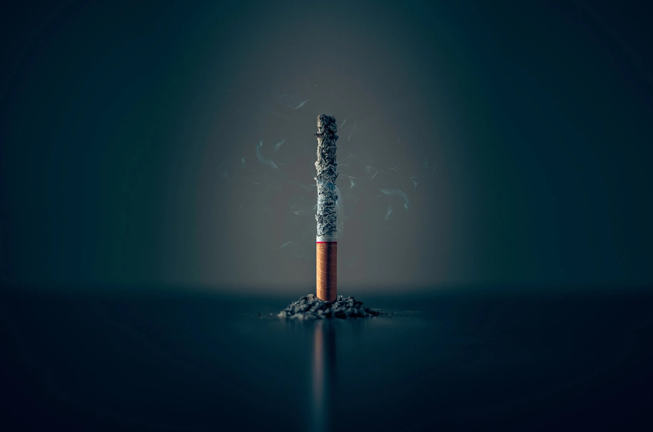 Lit cigarette standing vertically with ash on top and smoke curling around, set against a dark background.
