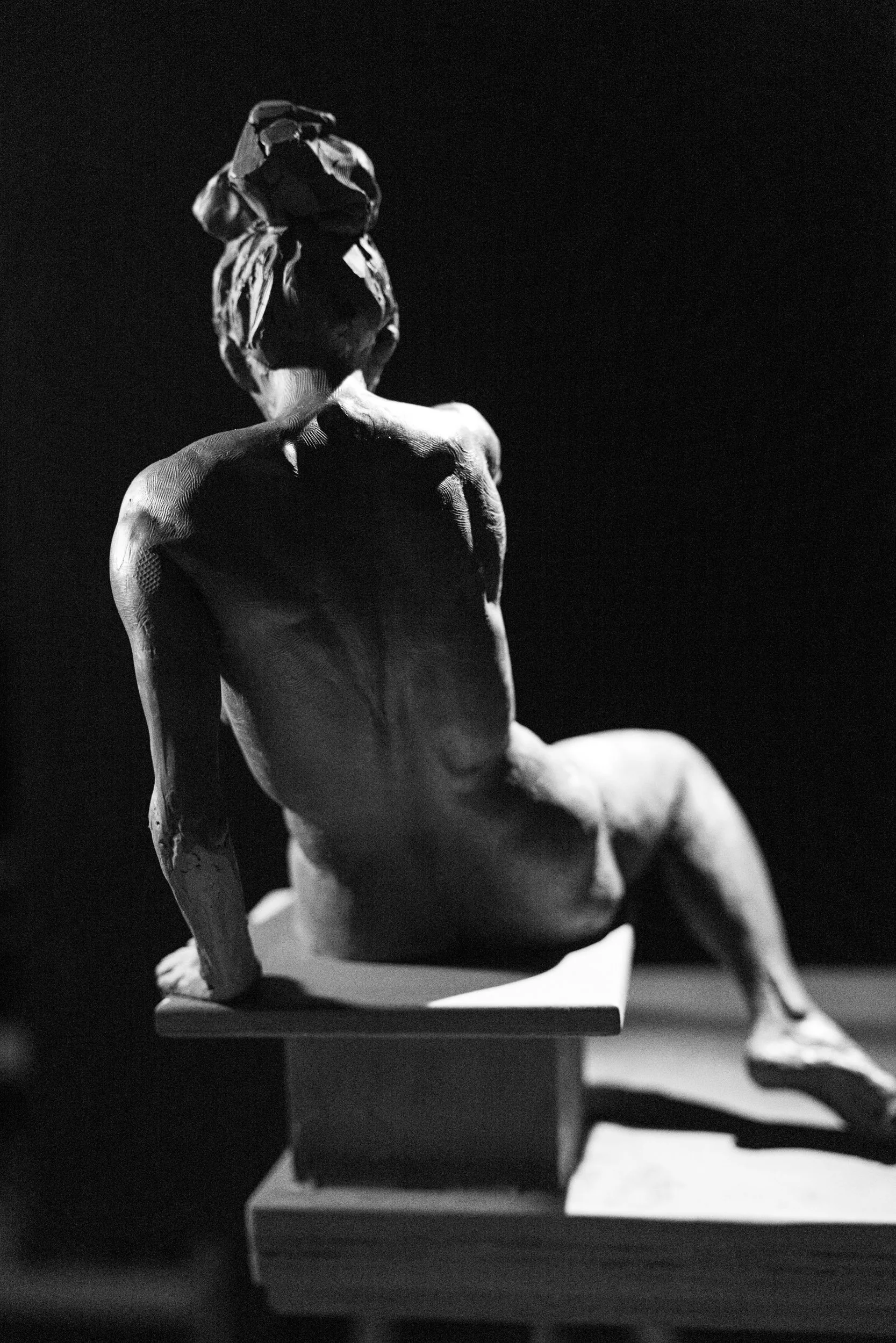 A black and white sculpture of a seated male figure, viewed from the back, with one hand raised to his head, displayed on a platform against a dark background.