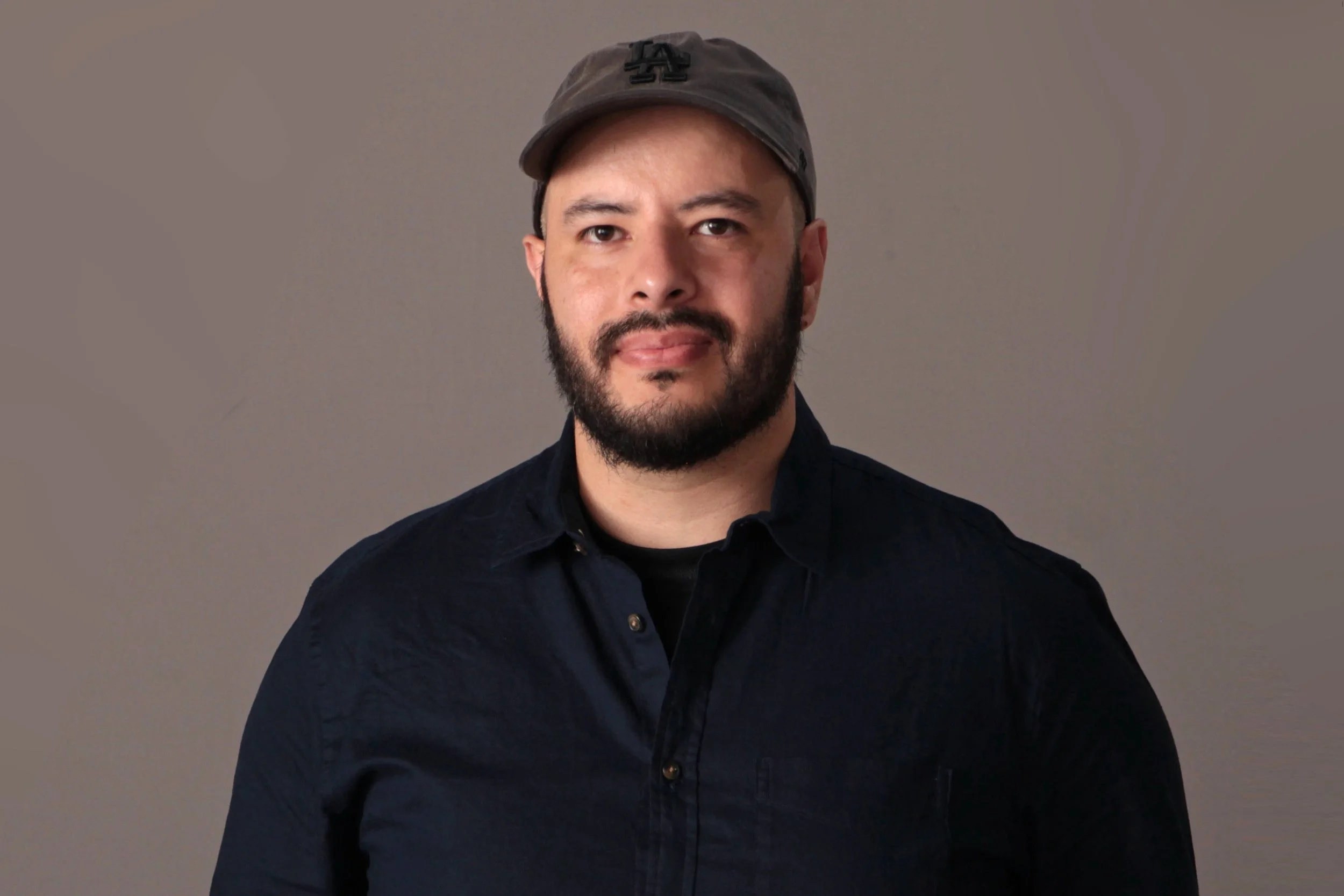 Portrait of Diego Olivares wearing a dark shirt and a gray cap, against a plain gray background.