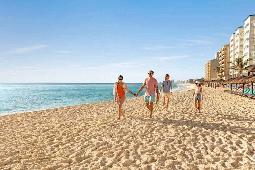 Two adults and two teenagers walk down a white sand beach with a turquoise ocean.