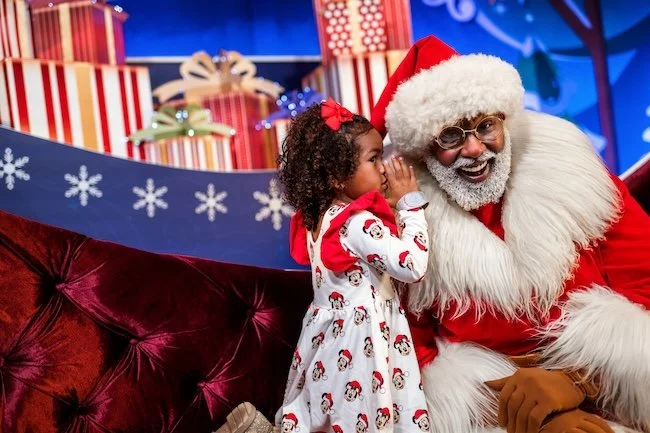 A little girl in a red and white dress with Mickey Mouse on it whispers in the ear of Santa Claus